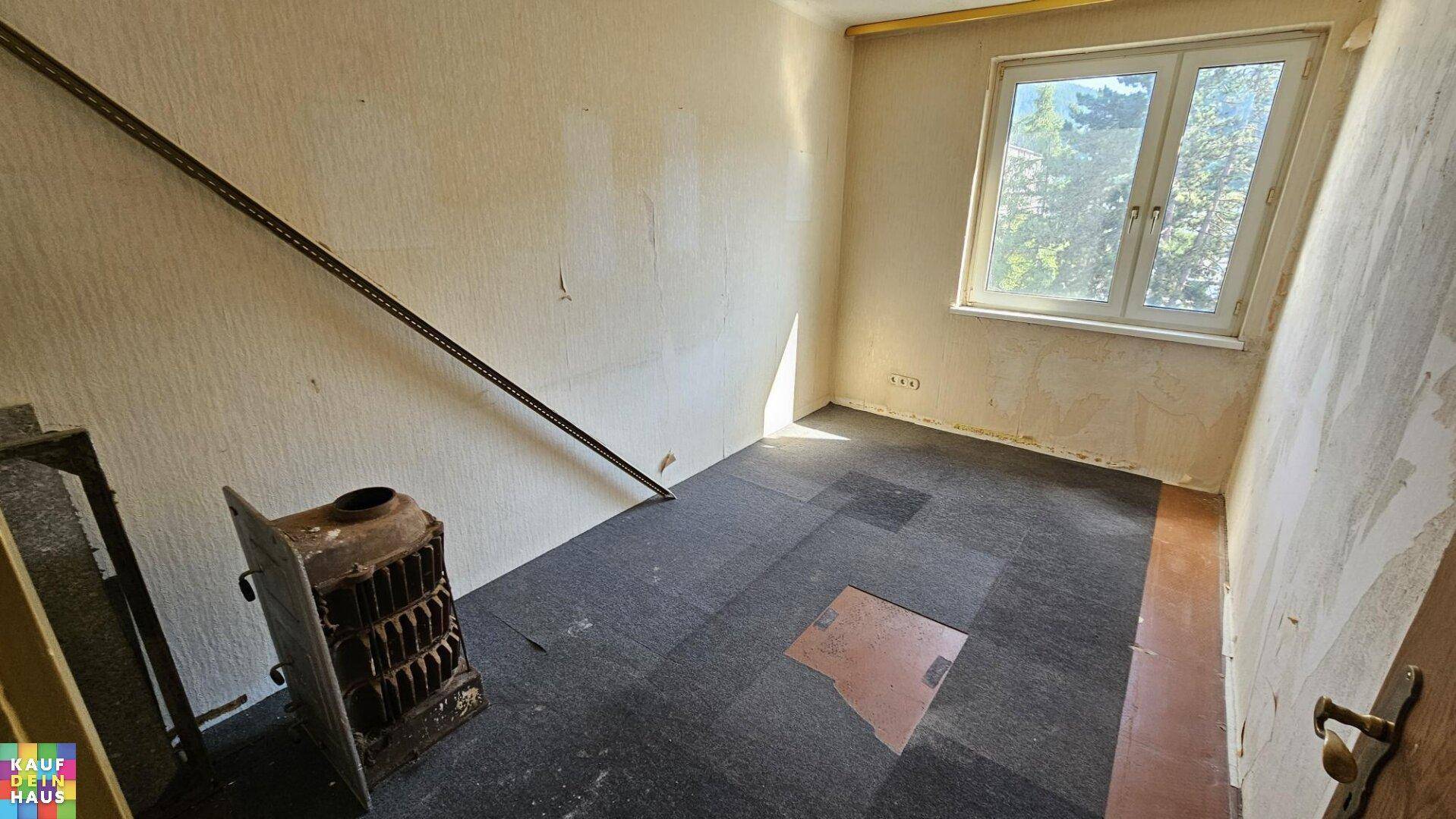 Renovierungsbedürftiges Zimmer mit altem Ofen und Teppichboden, bereit für eine Neugestaltung.