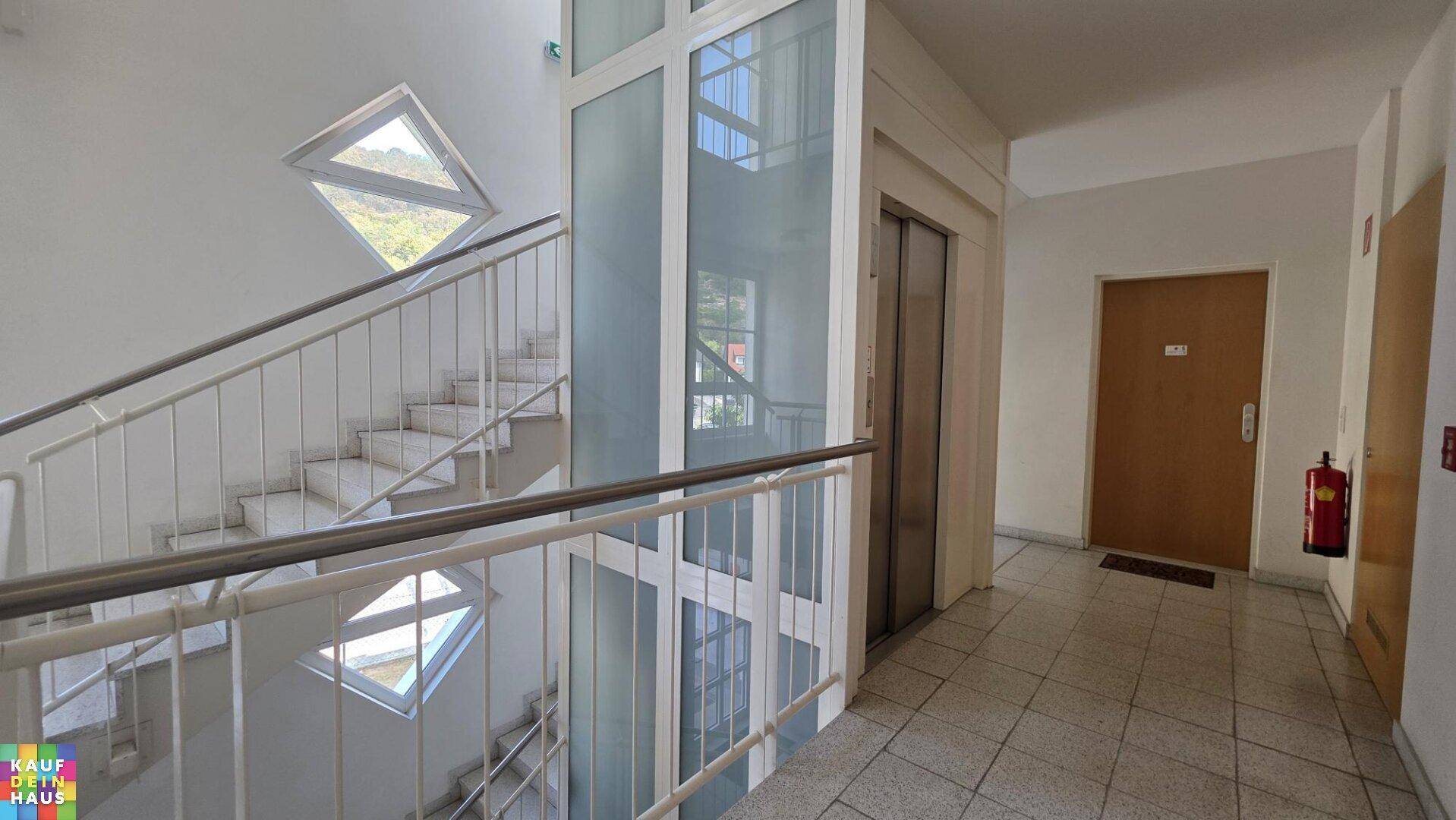Heller Treppenaufgang mit modernem Glaslift und Fliesenboden in einem Mehrfamilienhaus.