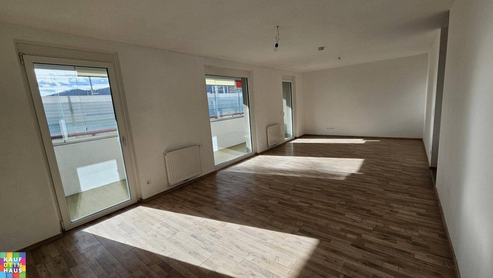 Geräumiges Wohnzimmer mit hellem Holzboden und großen Fenstern, die viel Tageslicht hereinlassen.