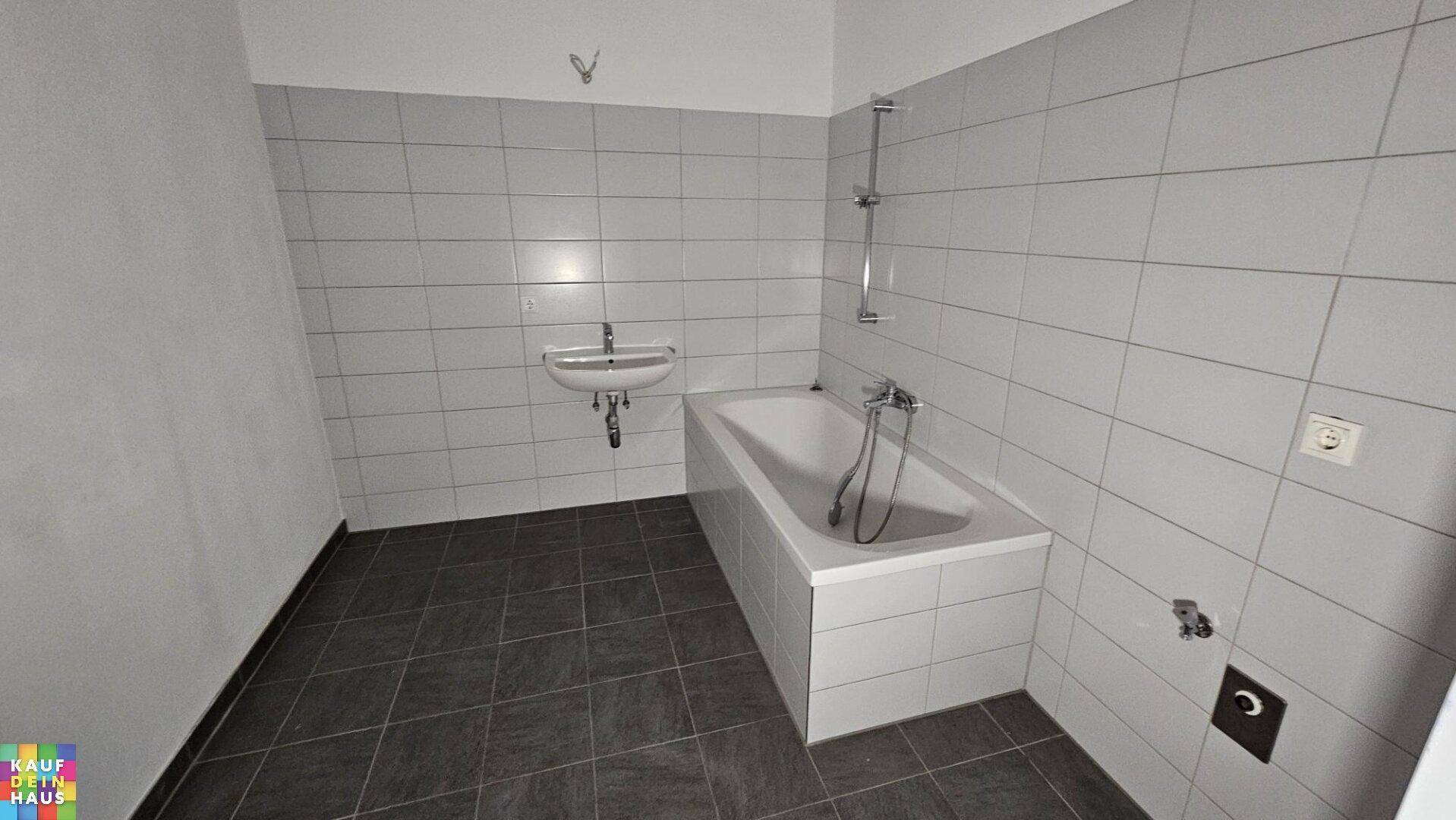 Funktionales Badezimmer mit weißen Wandfliesen, dunklem Boden und einer Badewanne mit Duschfunktion.