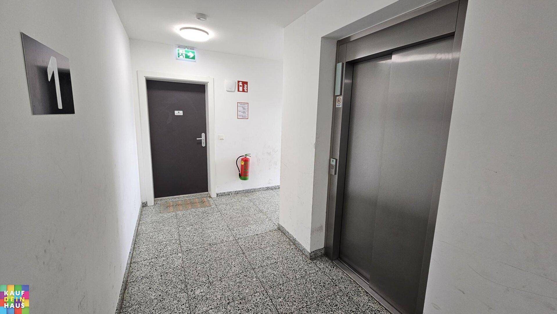 Gemeinschaftlicher Flur mit Aufzug und Fliesenboden, ausgestattet mit Brandschutzvorrichtungen.