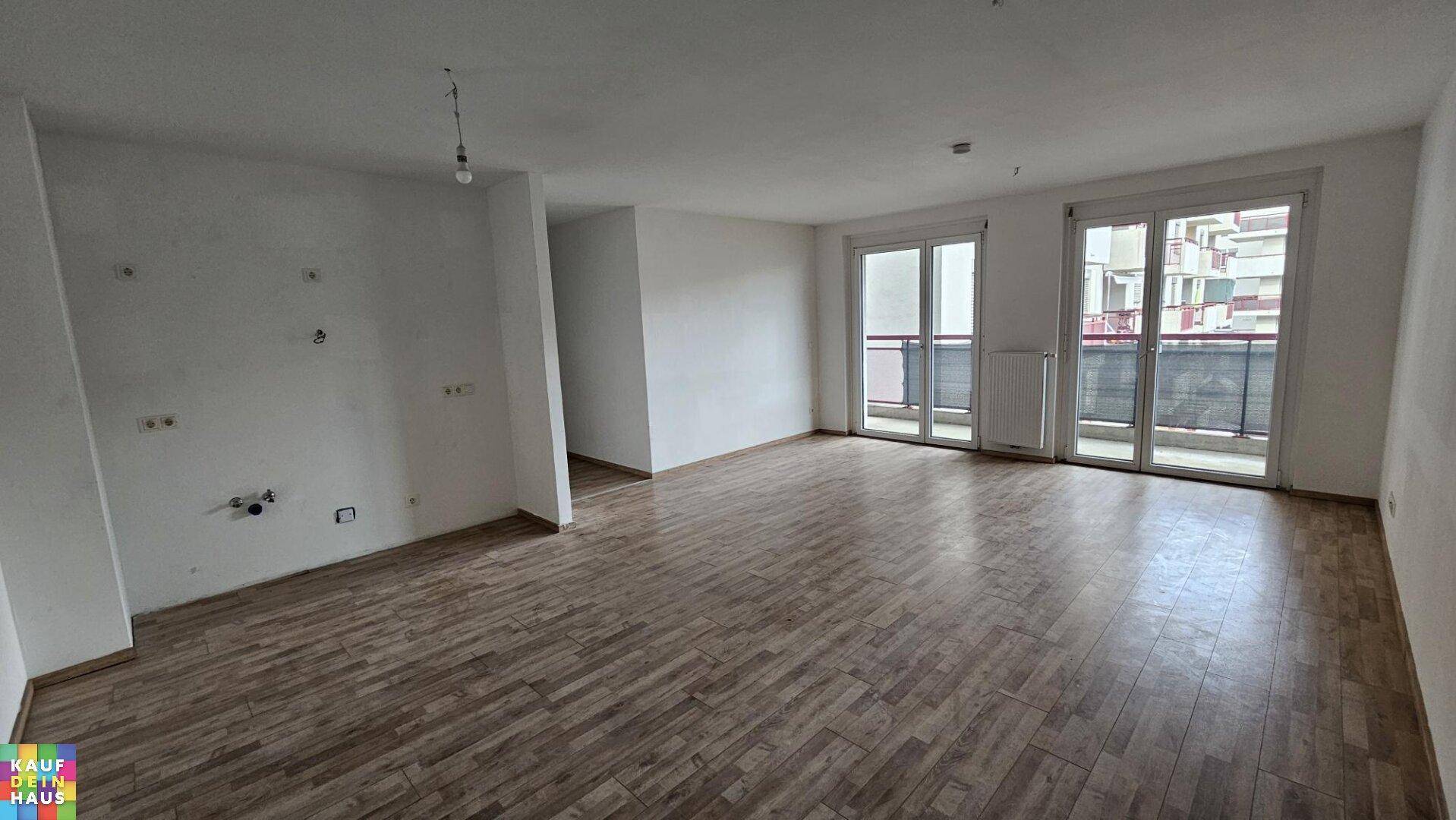 Helles, leeres Wohnzimmer mit Holzboden und Zugang zu einem Balkon.