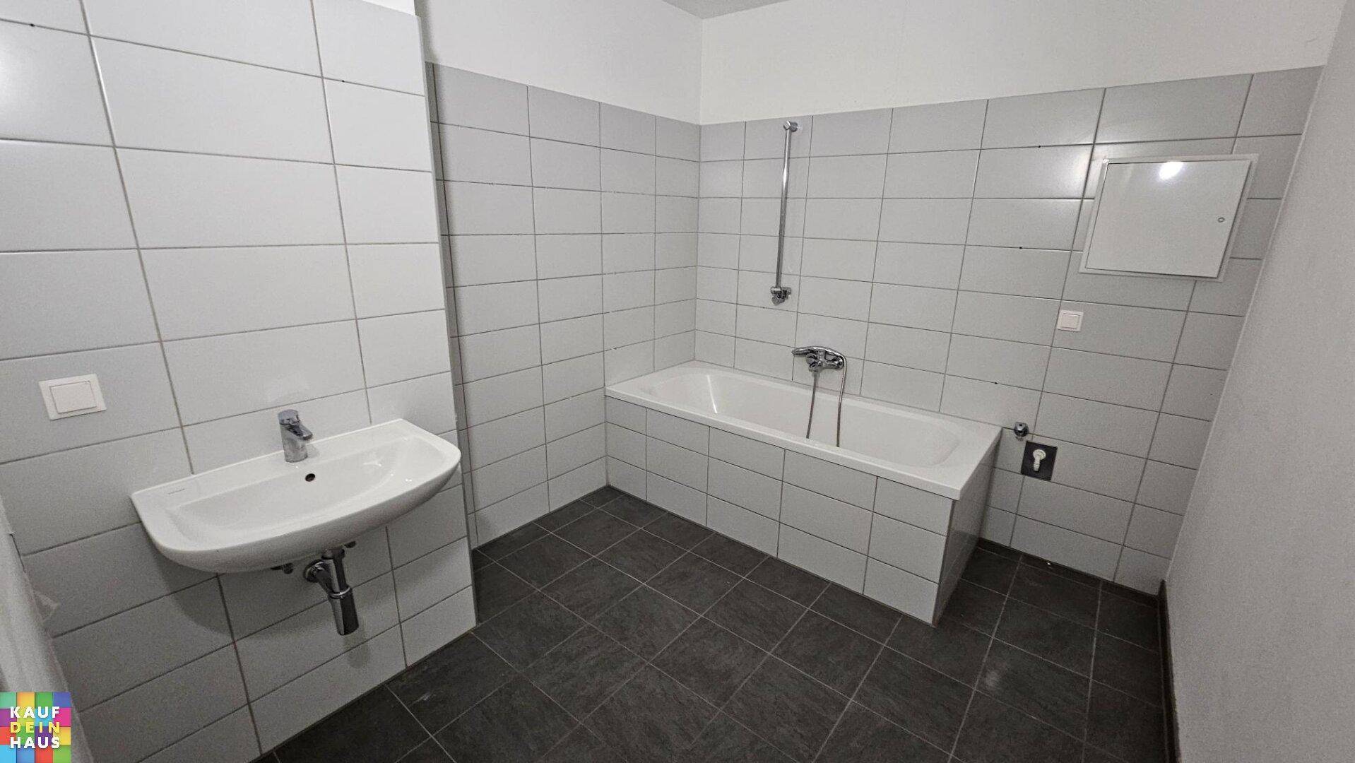 Badezimmer mit Badewanne, Waschbecken und hellgrauen Fliesen an Wänden und Boden.