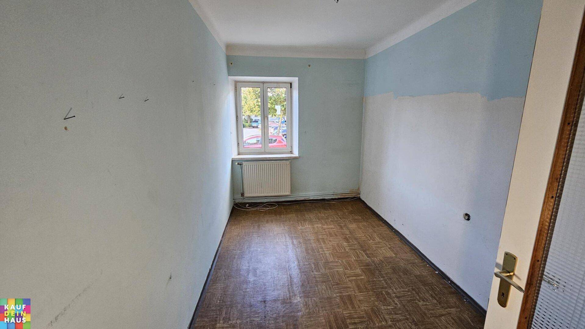 Leeres Zimmer mit zweifarbigen Wänden, Laminatboden und einem Fenster zur Straße.