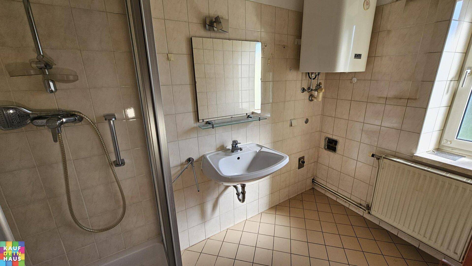 Badezimmer mit Dusche, Waschbecken und Boiler, komplett gefliest und mit Fenster ausgestattet.