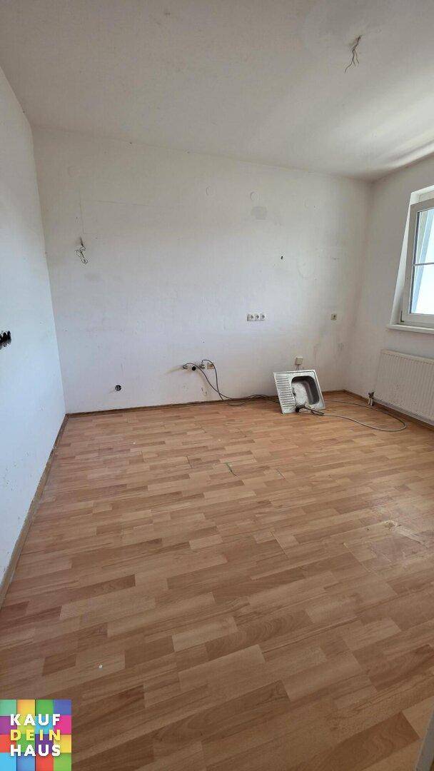 Leeres Zimmer mit hellem Laminatboden, weißen Wänden und einem Fenster mit Heizkörper.