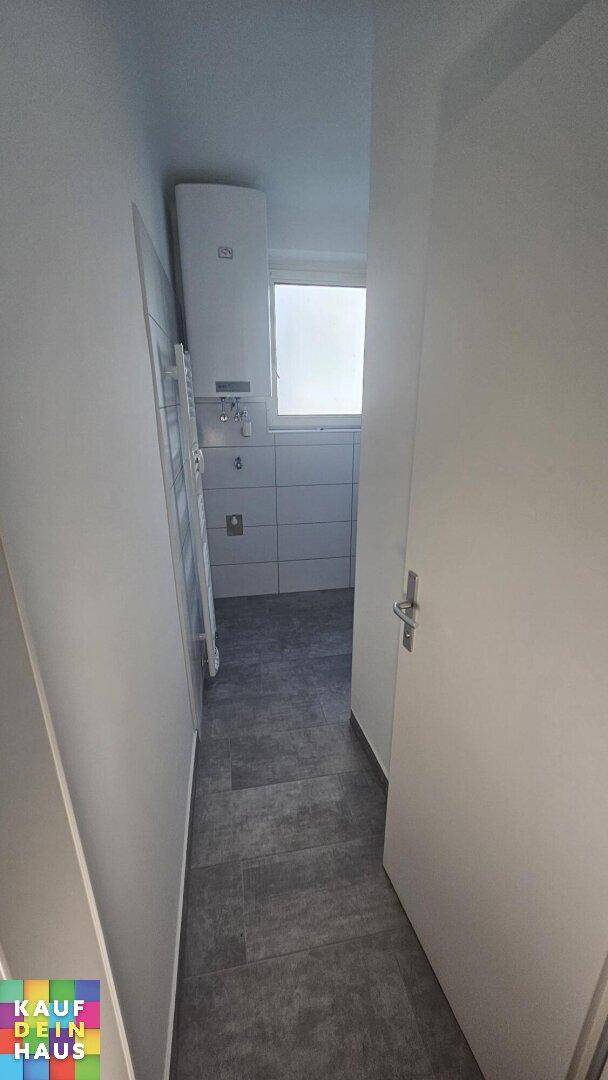Schmaler Flur mit grauen Fliesen, der zu einem Badezimmer oder Hauswirtschaftsraum führt.