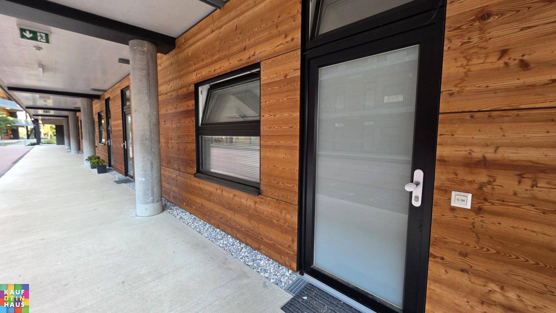 Eingangsbereich eines modernen Gebäudes mit Holzfassade und breitem Gehweg.