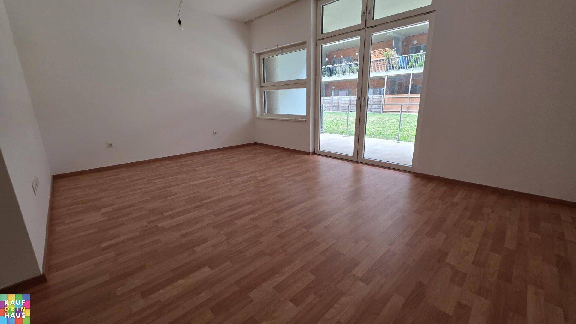 Heller, leerer Wohnraum mit Laminatboden und großen Fenstern zum Garten.
