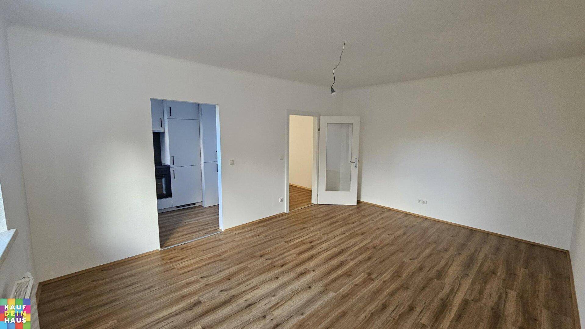 Geräumiger Wohnbereich mit Laminatboden, Zugang zur Küche und viel Tageslicht.