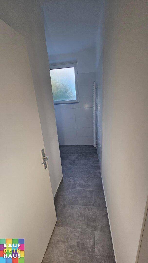 Schmaler Flur mit grauen Fliesen, der zu einem Badezimmer mit Fenster führt.