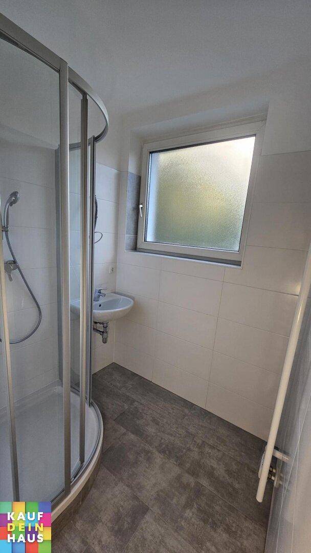 Badezimmer mit Eckdusche, Waschbecken, weißen Fliesen und einem Fenster für Tageslicht.