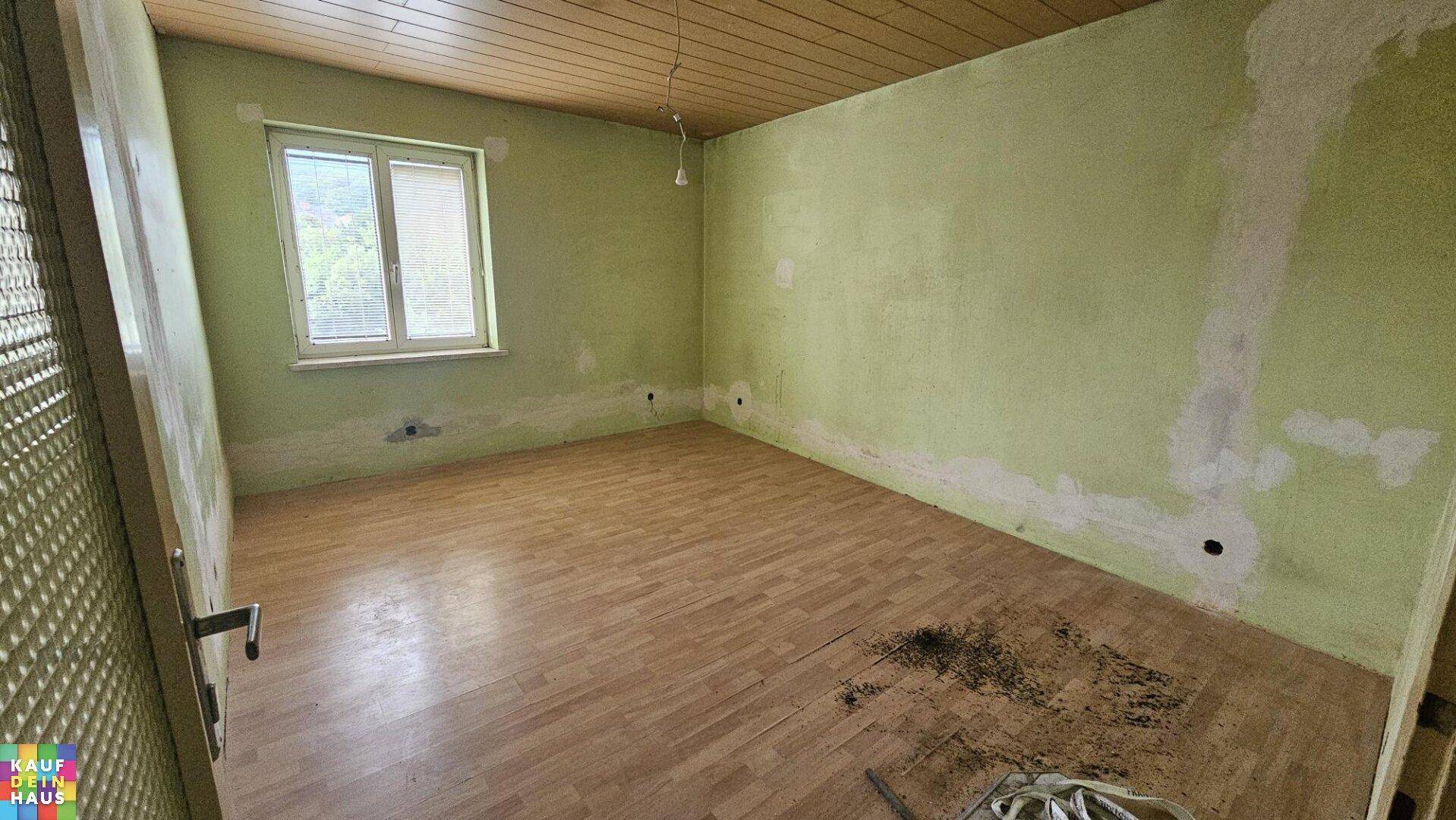 Leerer Raum mit teilweise gestrichenen grünen Wänden, Holzdeckenverkleidung und Laminatboden, bereit zur Renovierung.