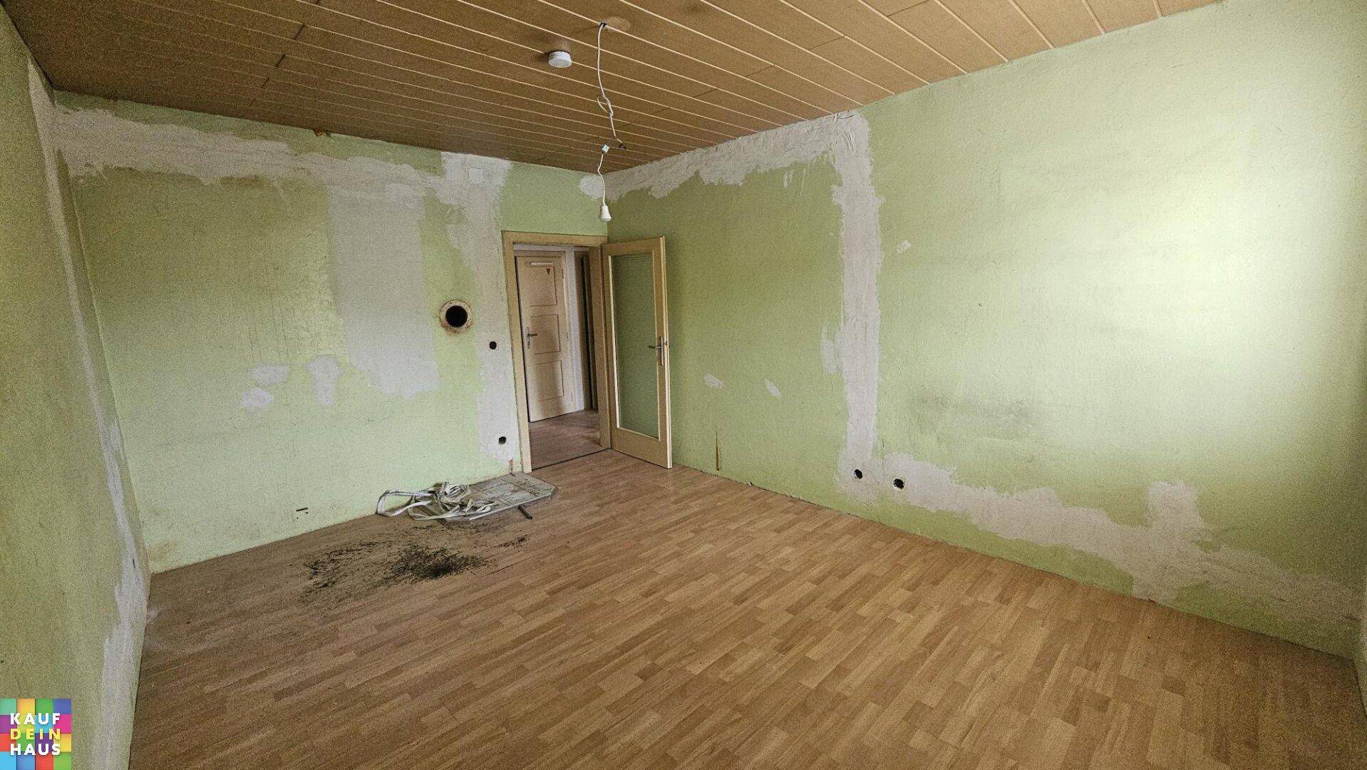 Leerer Raum mit Holzdeckenverkleidung und Laminatboden, teilweise gestrichenen Wänden und Zugang zu einem weiteren Zimmer.