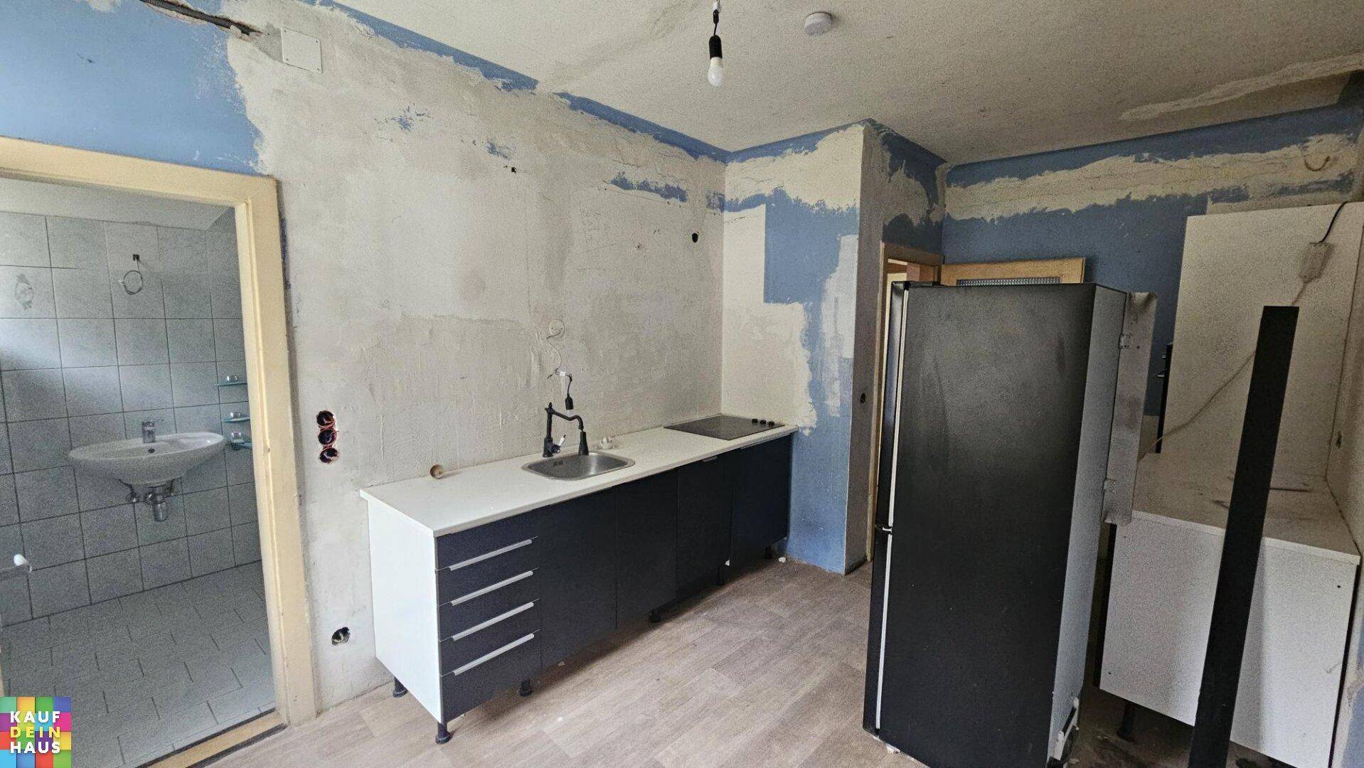 Küche in Renovierung mit Spüle, Kochfeld, dunklen Unterschränken und Kühlschrank, angrenzend an ein Badezimmer.