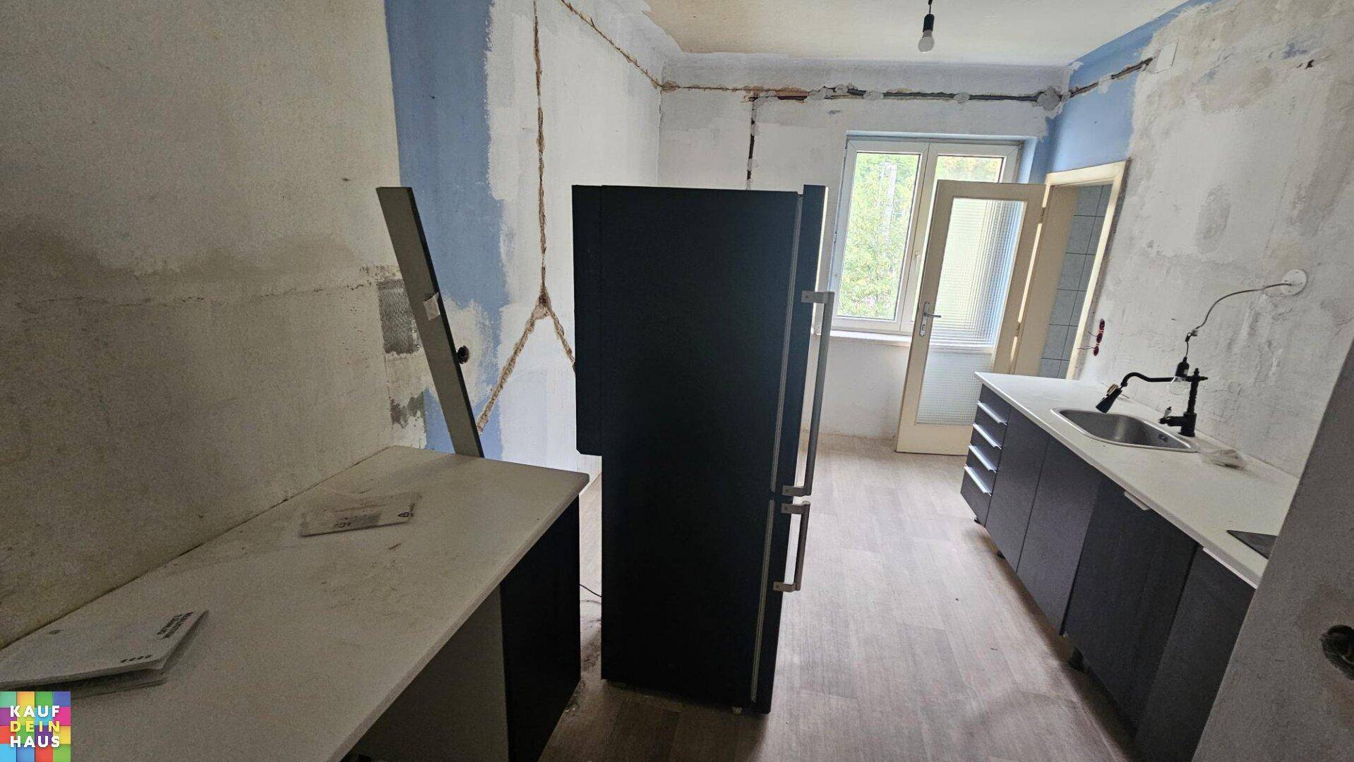 Küchenbereich in Renovierung mit schwarzem Kühlschrank, Arbeitsplatte und Fenster, mit Blick ins Grüne.