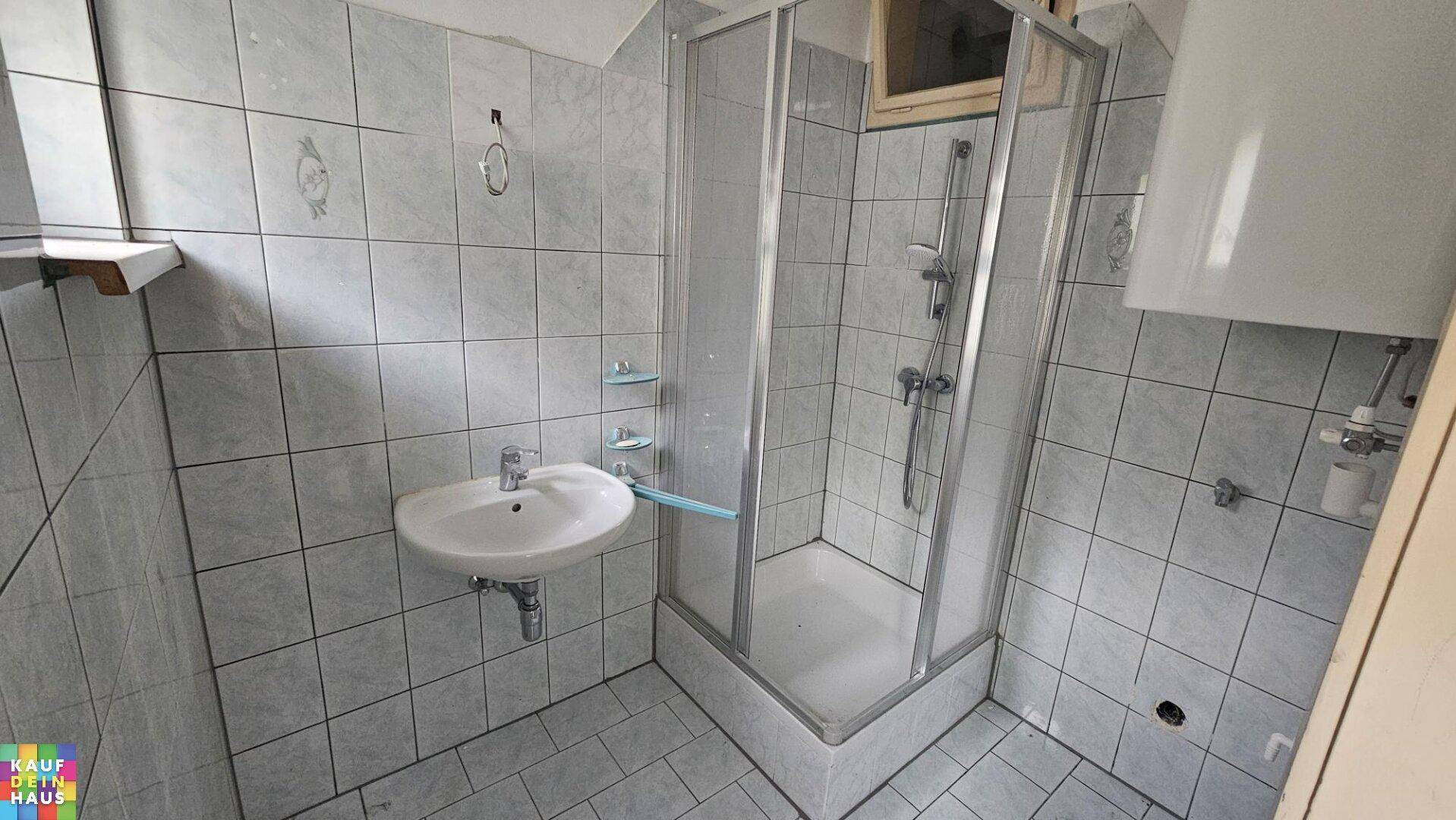 Gefliestes Badezimmer mit Dusche, Waschbecken und Fenster, in einem älteren, aber funktionalen Zustand.