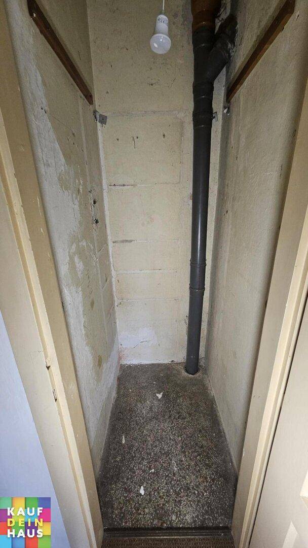 Schmaler Abstellraum mit Betonwänden, sichtbarer Rohrleitung und einfachem Bodenbelag.