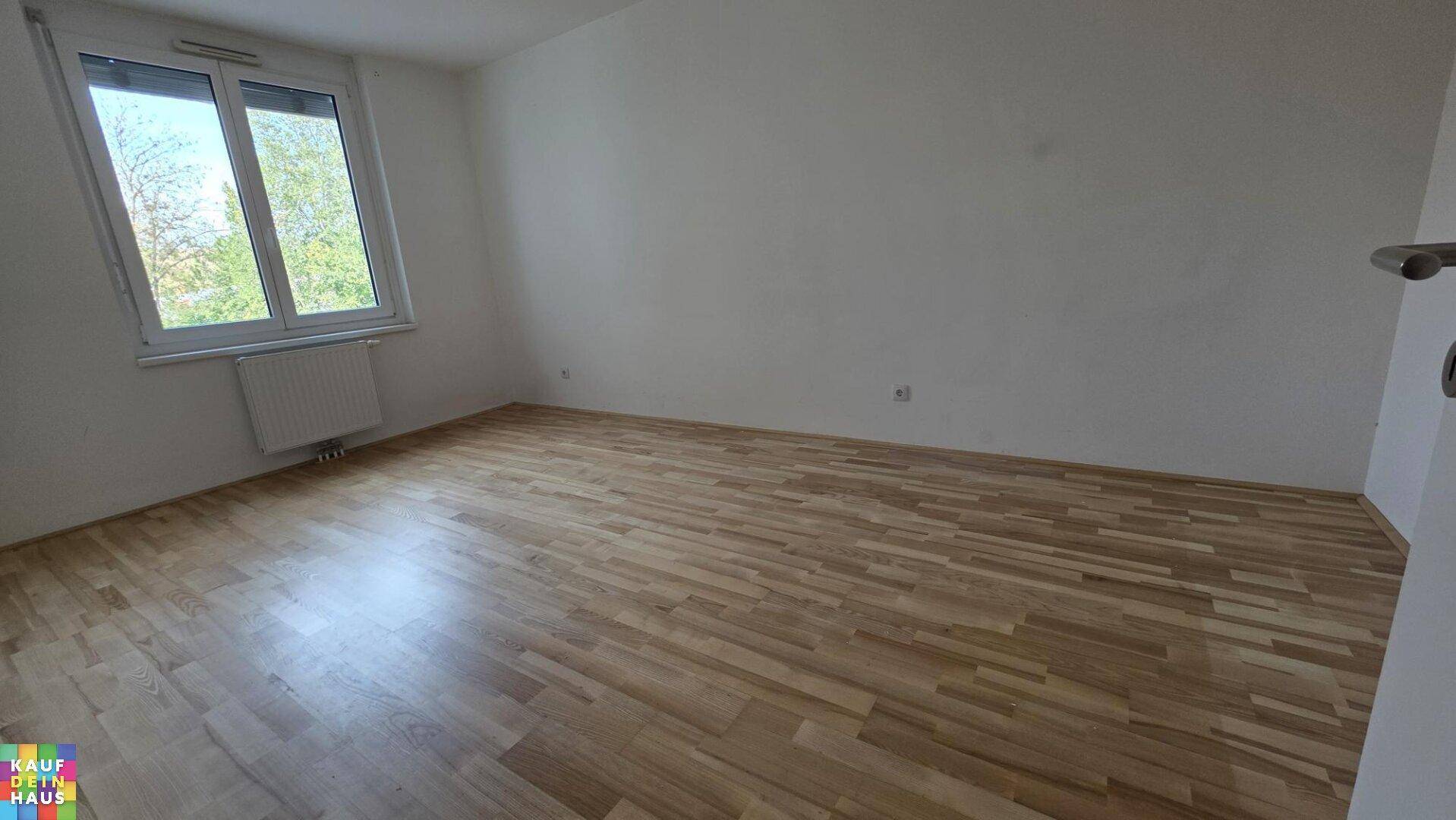 Leeres Schlafzimmer mit Holzparkett und großem Fenster, das viel Tageslicht hereinlässt.