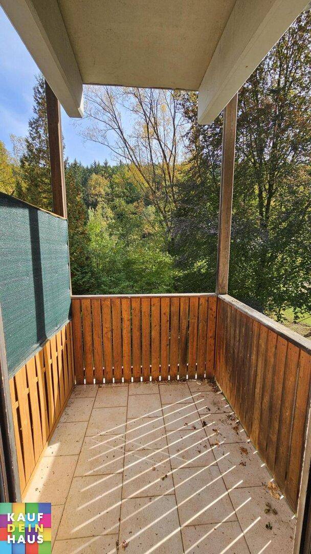 Sonniger Balkon mit Fliesenboden und Holzgeländer, umgeben von grünen Bäumen.