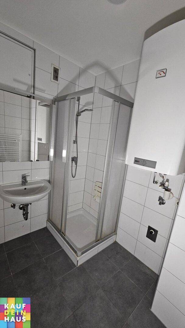 Funktionales Badezimmer mit weißen Fliesen, Duschkabine, Waschbecken und Warmwasserboiler.