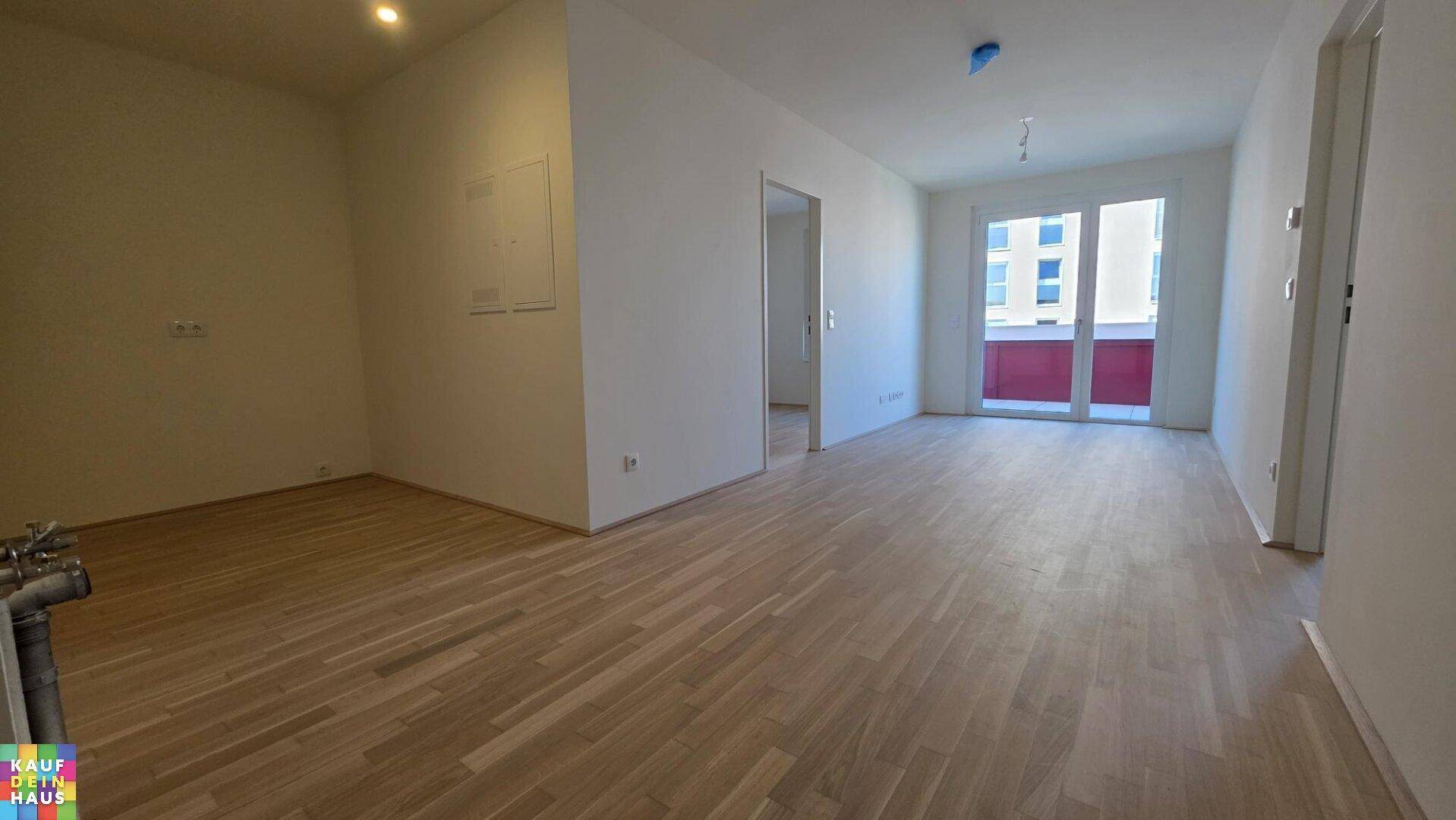 Geräumiger, leerer Wohnraum mit hellem Laminatboden, weißen Wänden und Zugang zum Balkon.