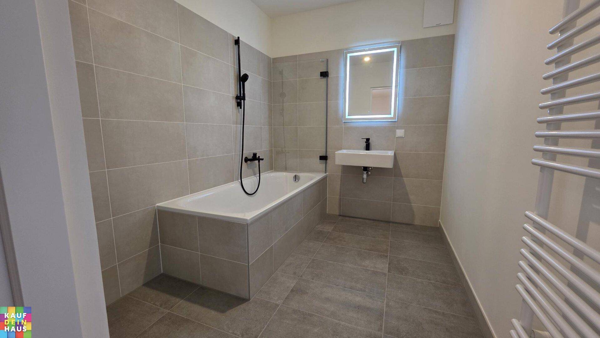 Modernes Badezimmer mit Badewanne, beleuchtetem Spiegel, Einzelwaschbecken und Handtuchheizkörper.