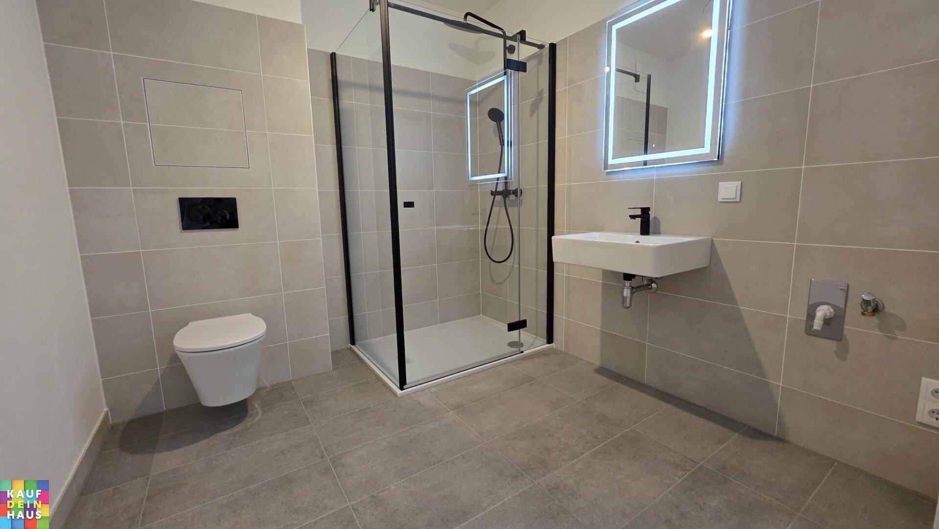 Modernes Badezimmer mit grauen Fliesen, ebenerdiger Dusche und beleuchtetem Spiegel.