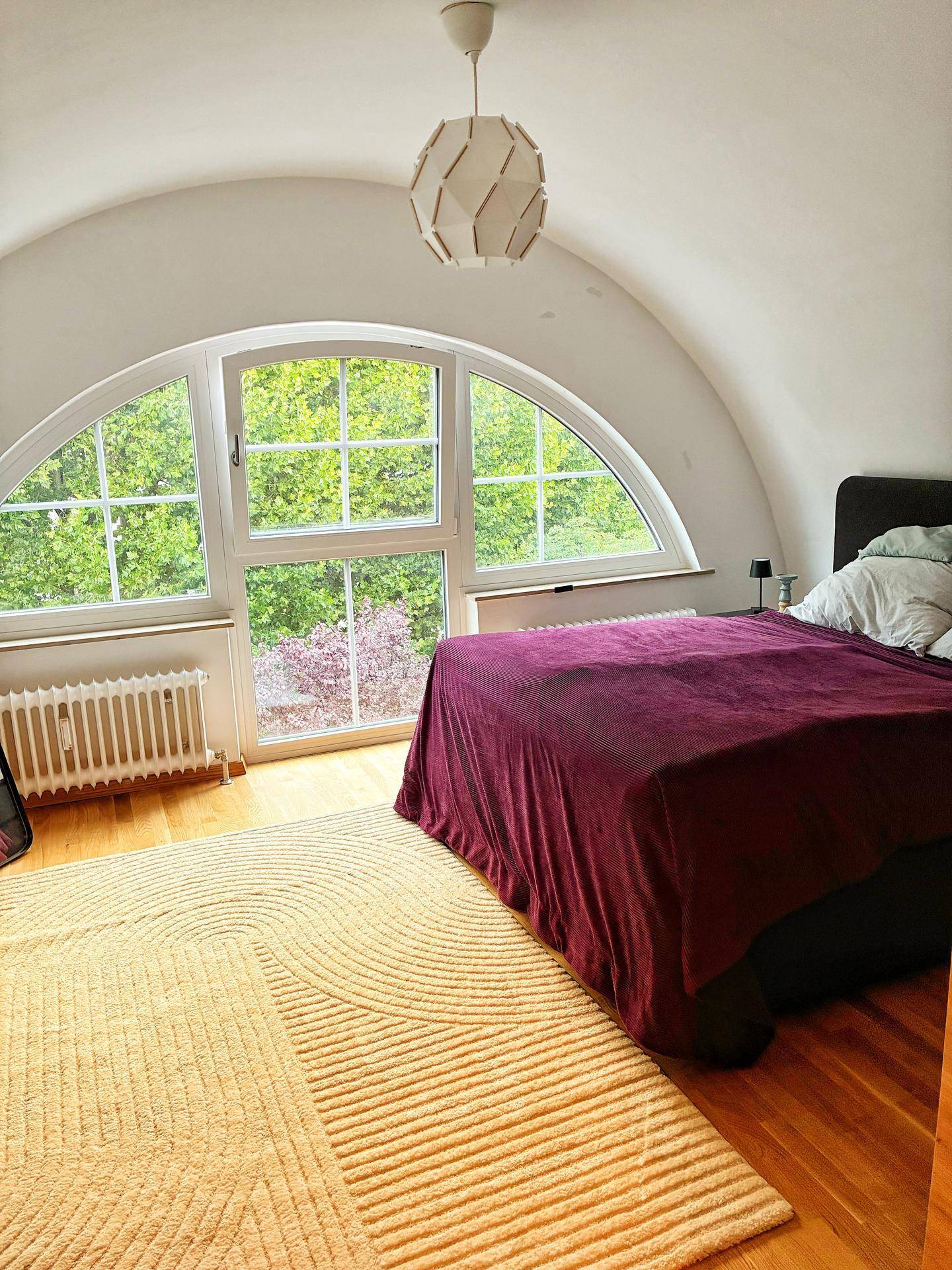 Schlafzimmer mit großem Rundbogenfenster, Holzboden und Blick ins Grüne, unter einer gewölbten Decke.