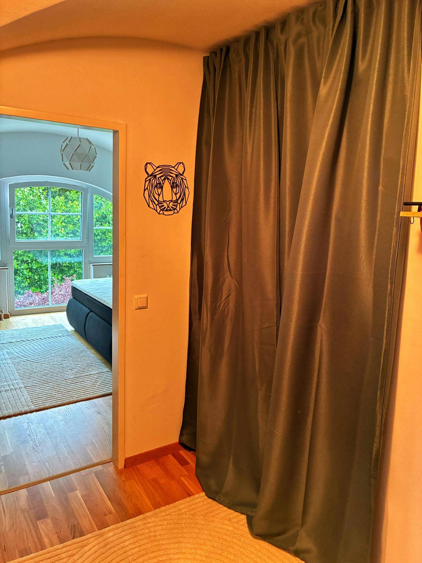 Flur mit Vorhang und Blick in ein helles Schlafzimmer mit großen Fenstern und viel Grünblick.