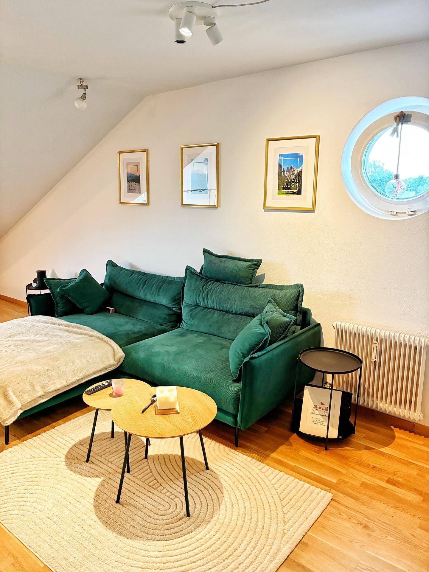 Gemütliches Wohnzimmer mit grüner Couch, Holzboden und rundem Fenster in der Dachschräge für viel Licht.