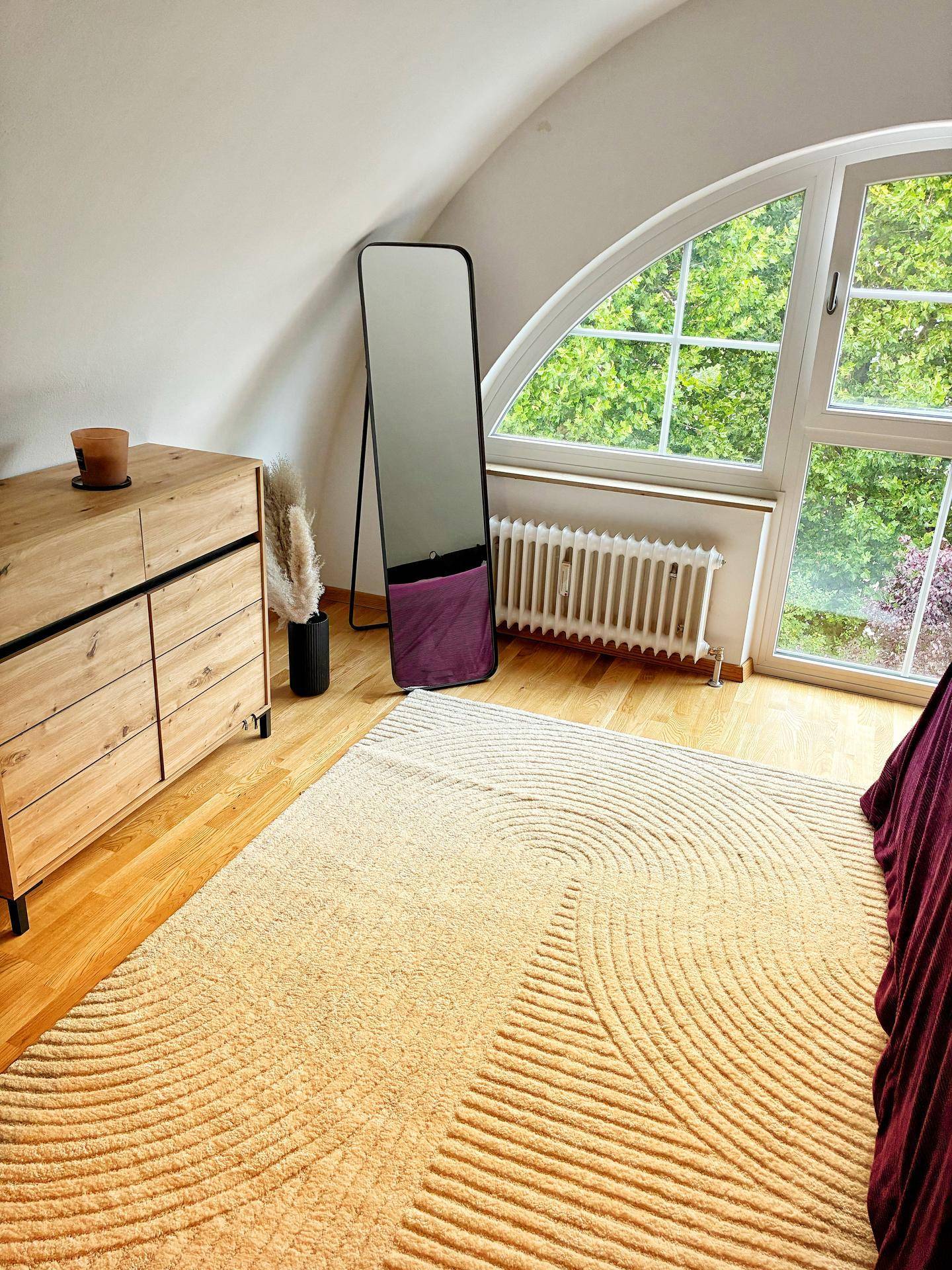 Detailansicht des Schlafzimmers mit Holzkommode, großem Spiegel und Rundbogenfenster mit Grünblick.