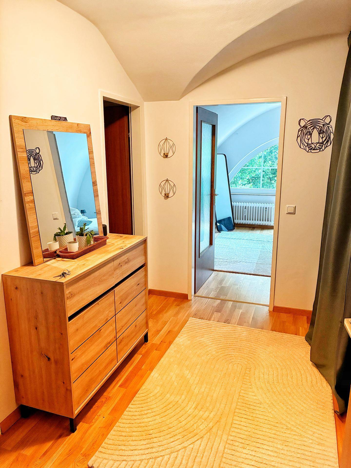 Heller Flur mit Holzkommode, Spiegel und Zugang zu einem Schlafzimmer mit großen Fenstern.
