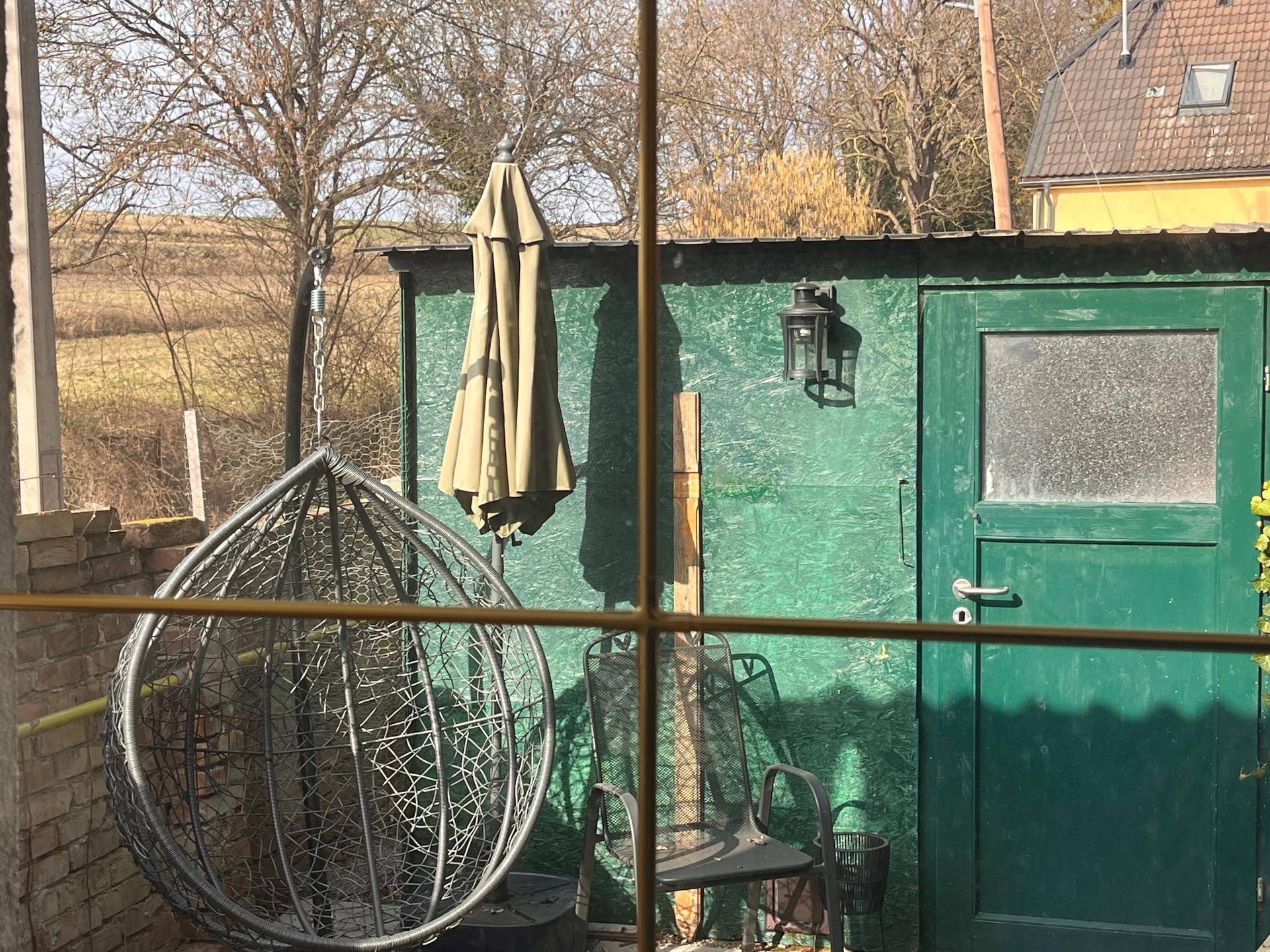 Blick aus dem Fenster auf einen Gartenbereich mit Schaukelstuhl und Geräteschuppen.