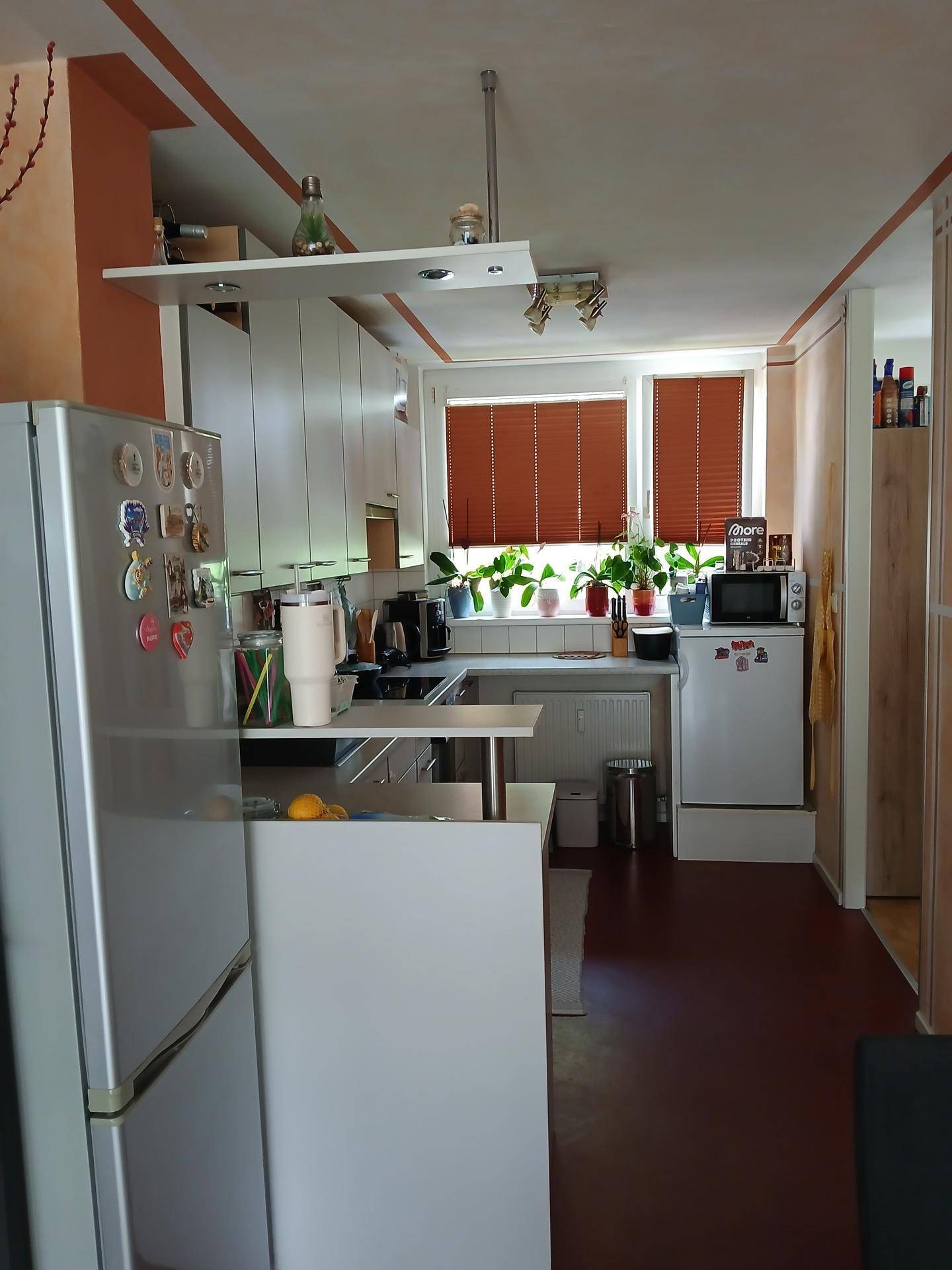 Funktionale Küche mit weißen Schränken, silbernem Kühlschrank und Pflanzen auf der Fensterbank.