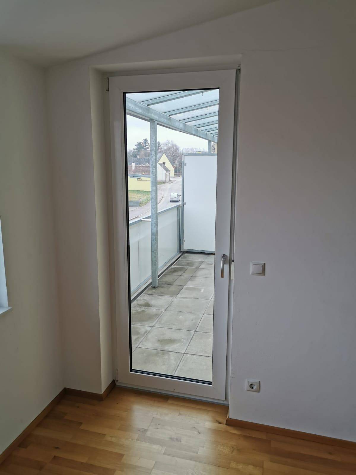 Heller Raum mit Holzboden und Zugang zu einem überdachten Balkon durch eine Glastür.