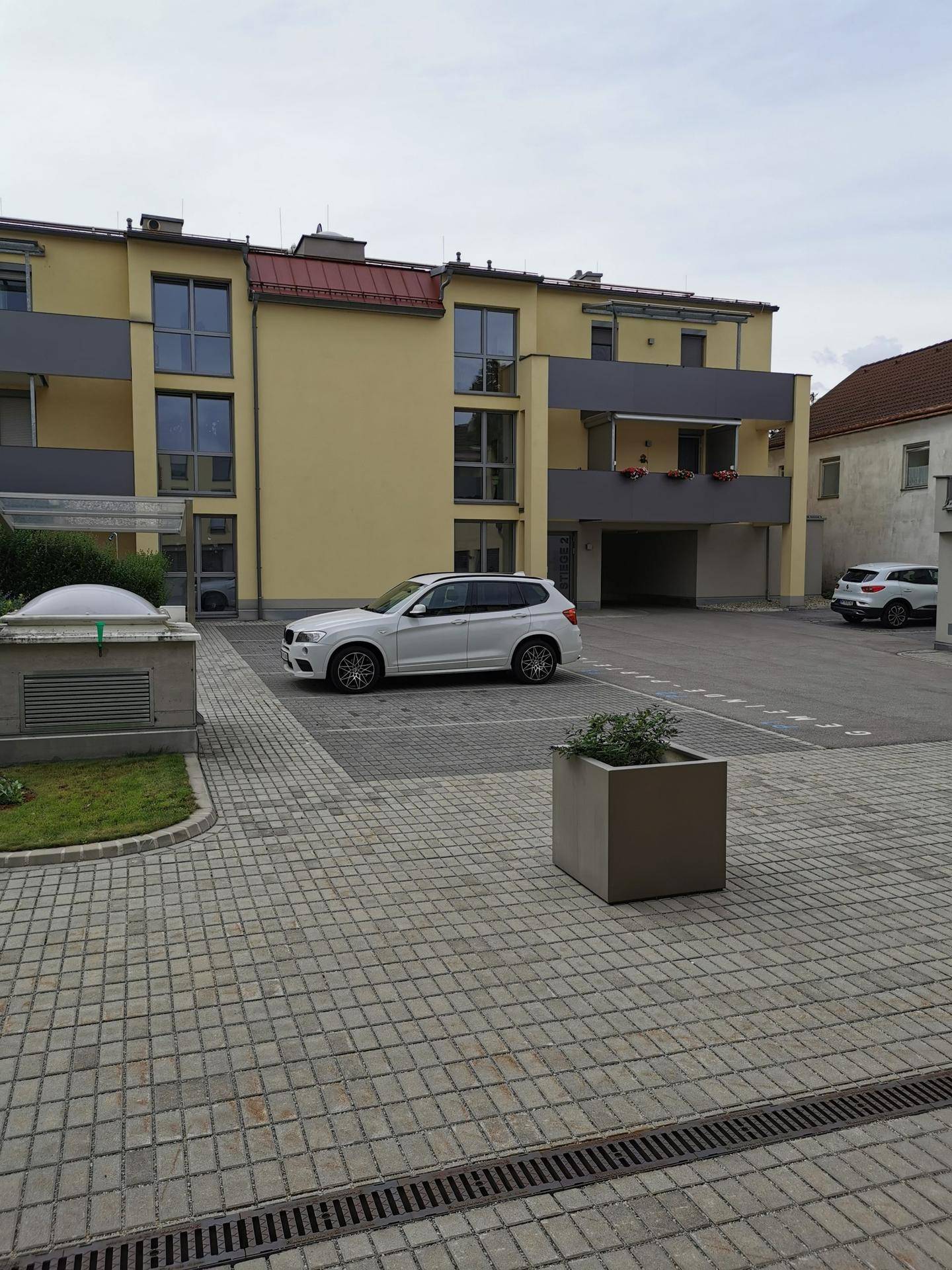 Moderne Gebäudefassade mit Balkonen und großen Fenstern, davor ein gepflasterter Parkplatz.