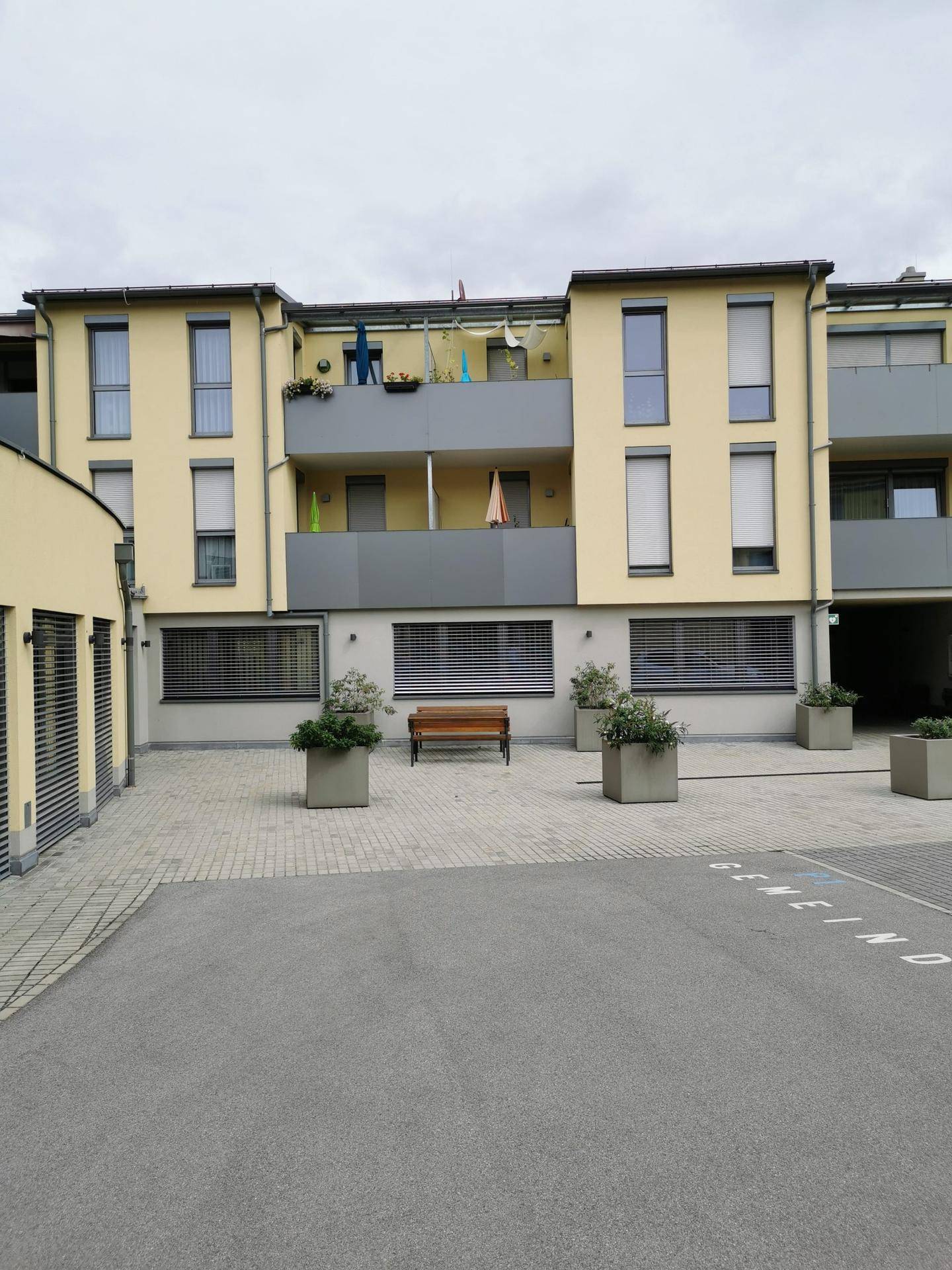 Rückansicht eines modernen Wohngebäudes mit Balkonen und einem gepflasterten Innenhof mit Bänken.