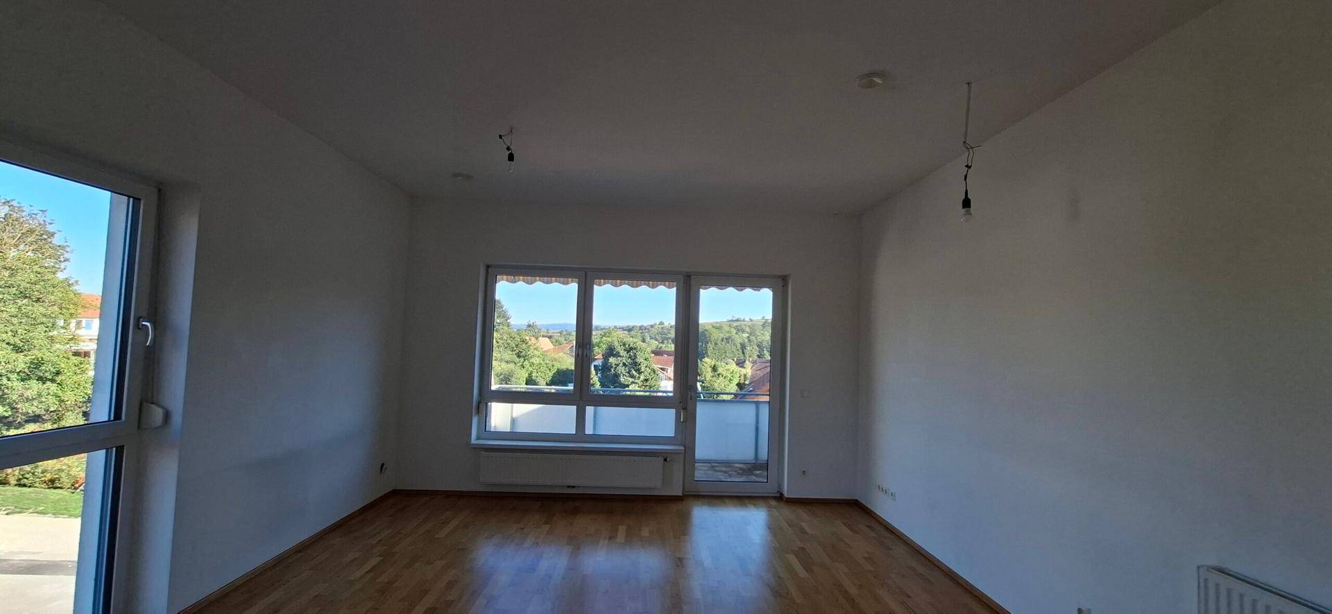Heller Wohnbereich mit Holzboden und weitem Ausblick durch große Fenster und Balkontür.