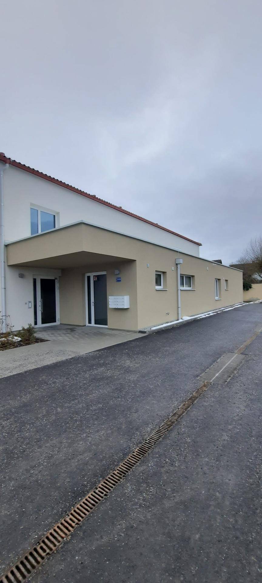 Seitenansicht eines Gebäudes mit heller Fassade, rotem Ziegeldach und gepflastertem Zugangsbereich.