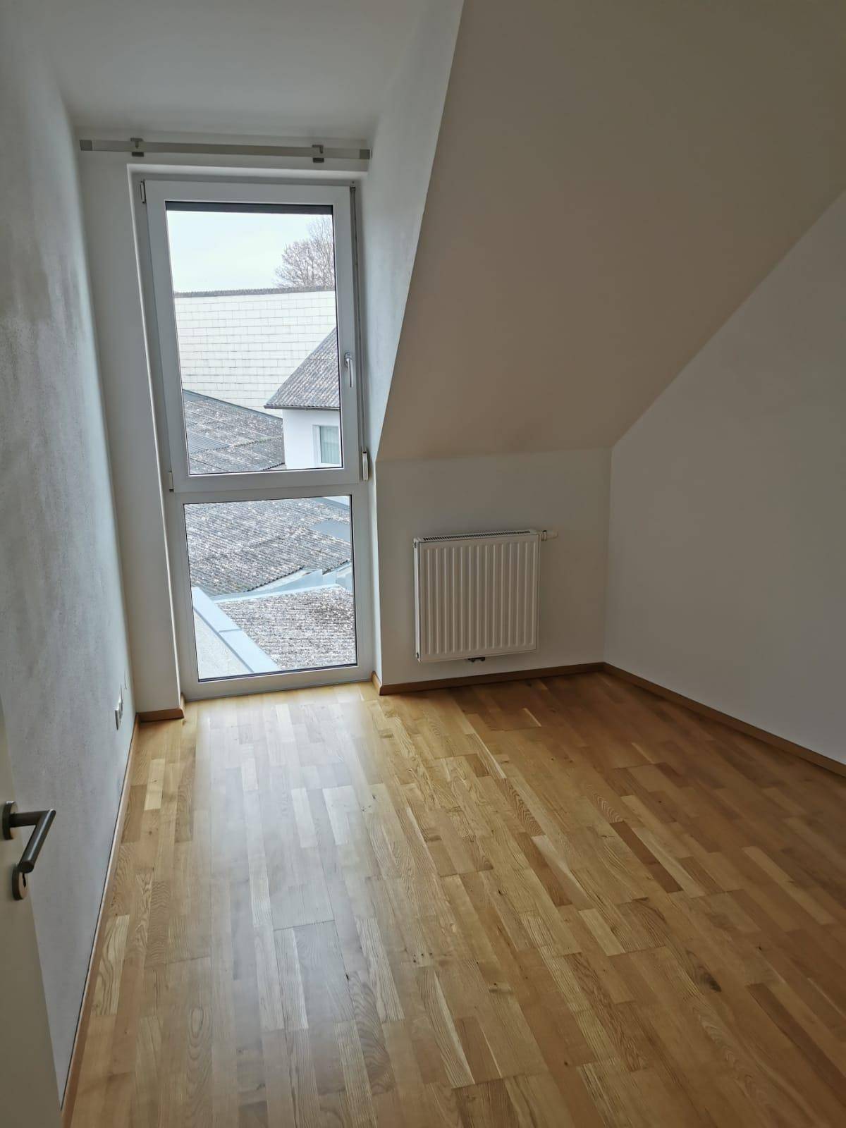 Leerer Raum mit Holzboden, schräger Decke und hohem Fenster, das einen Blick nach draußen bietet.