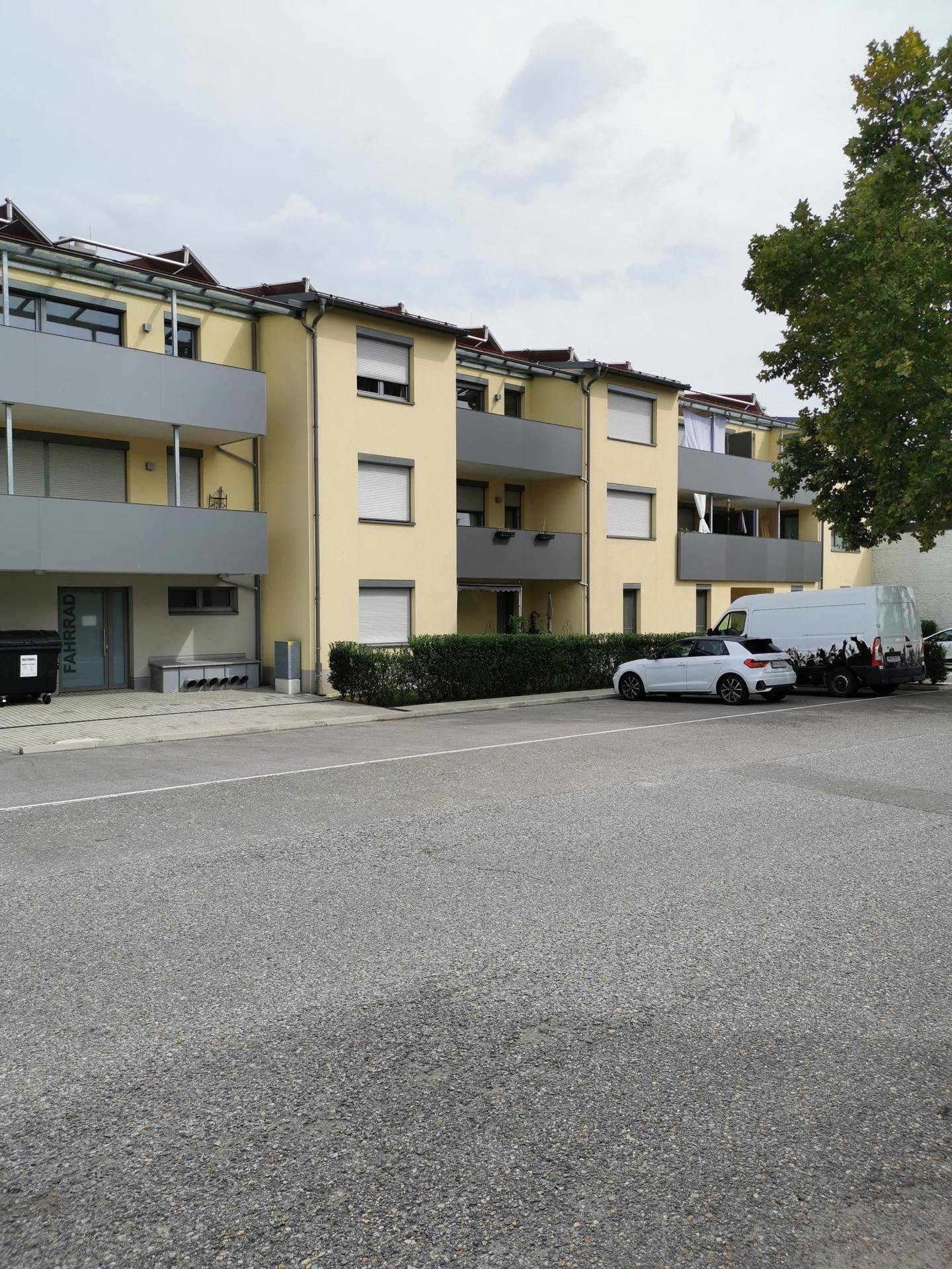Moderne Wohnanlage mit Balkonen und einem Parkplatz, umgeben von Grünflächen und Bäumen.