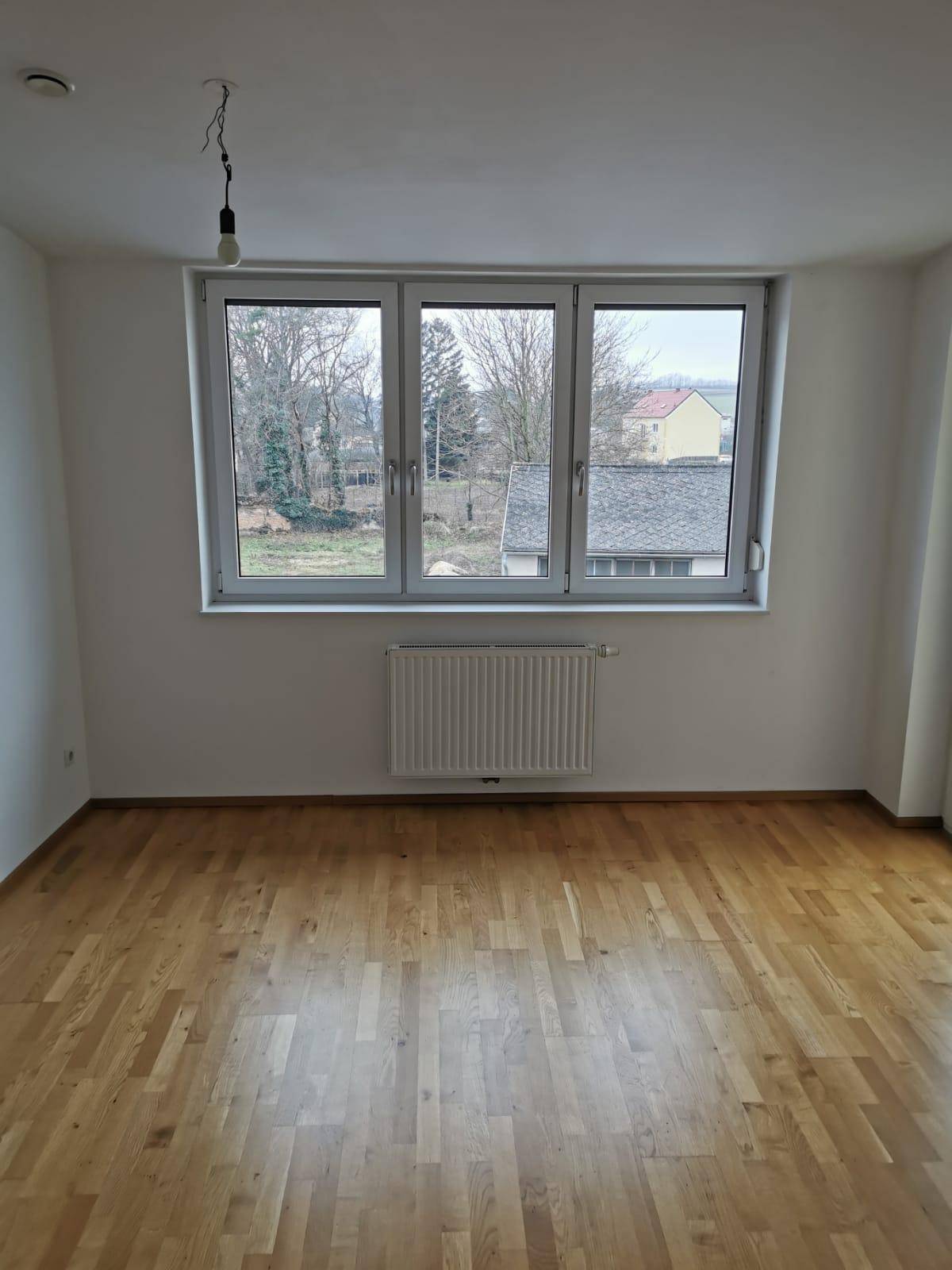 Leerer Raum mit Holzboden, weißen Wänden und einem großen Fenster mit Blick ins Grüne.