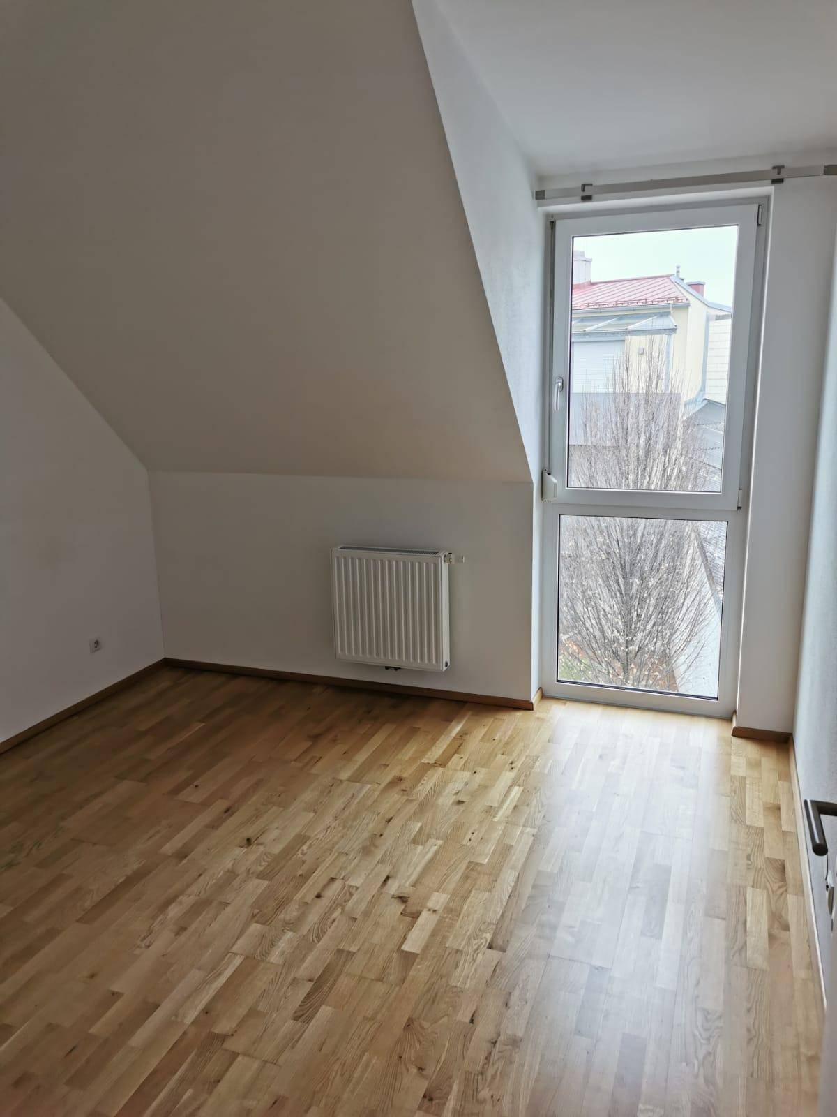 Heller Raum mit Holzboden, schräger Decke und einem hohen Fenster, das viel Licht hereinlässt.