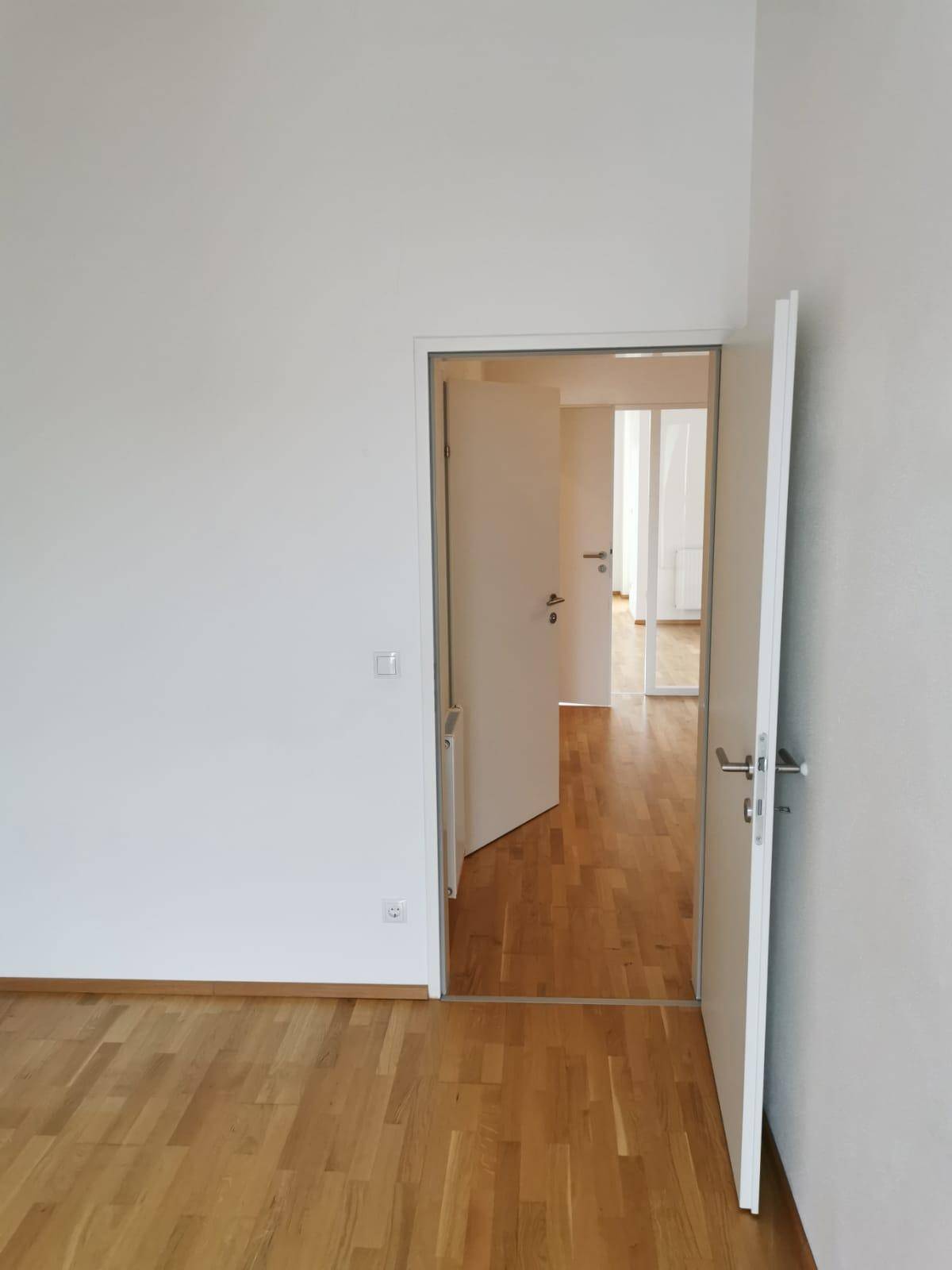 Heller Raum mit Holzboden und offener Tür, die den Blick auf einen weiteren Raum freigibt.