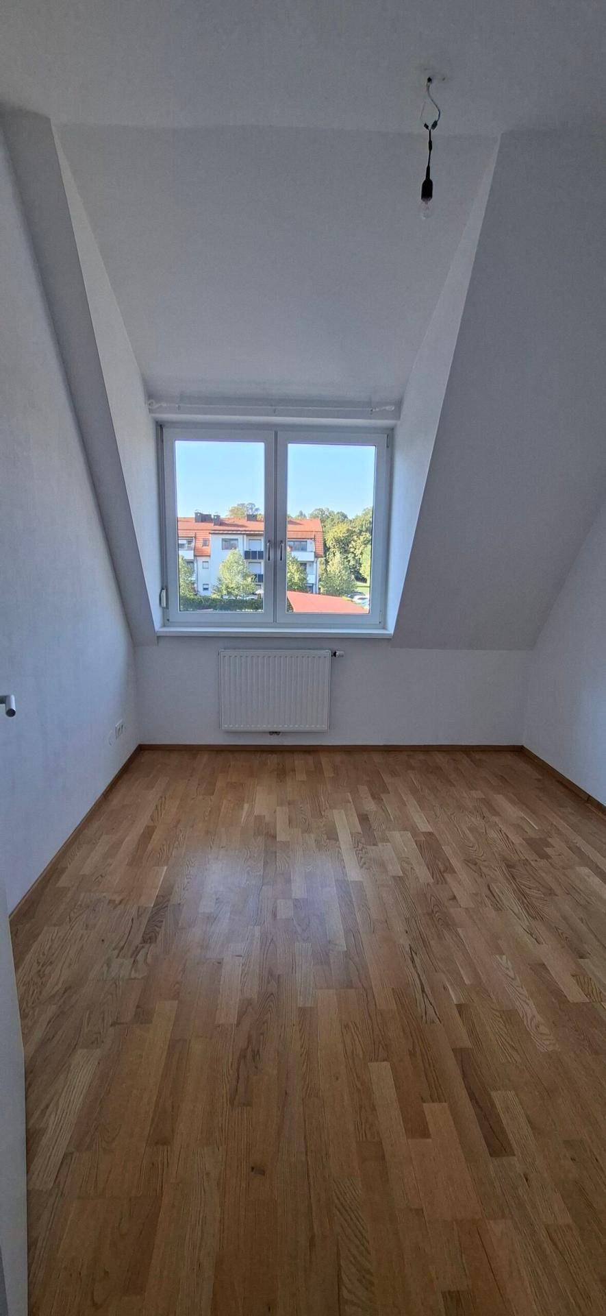 Helles Schlafzimmer im Dachgeschoss mit Holzboden und schrägen Wänden unter einem Fenster.