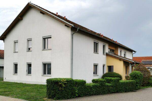 Seitenansicht eines Mehrfamilienhauses mit weißer Fassade, gelbem Anbau und einer gepflegten Hecke.