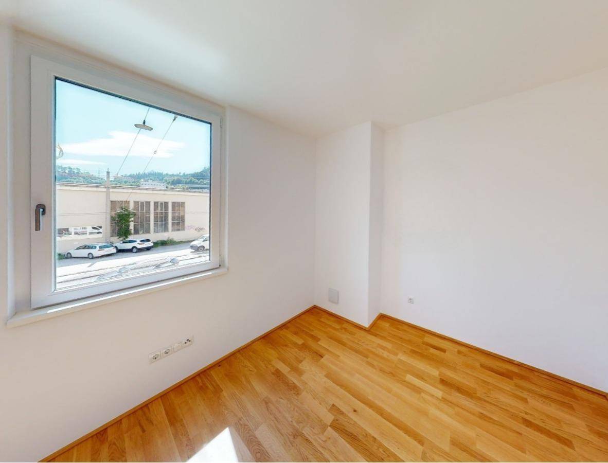 Helles, leeres Zimmer mit Holzboden und großem Fenster mit Blick auf die Straße.