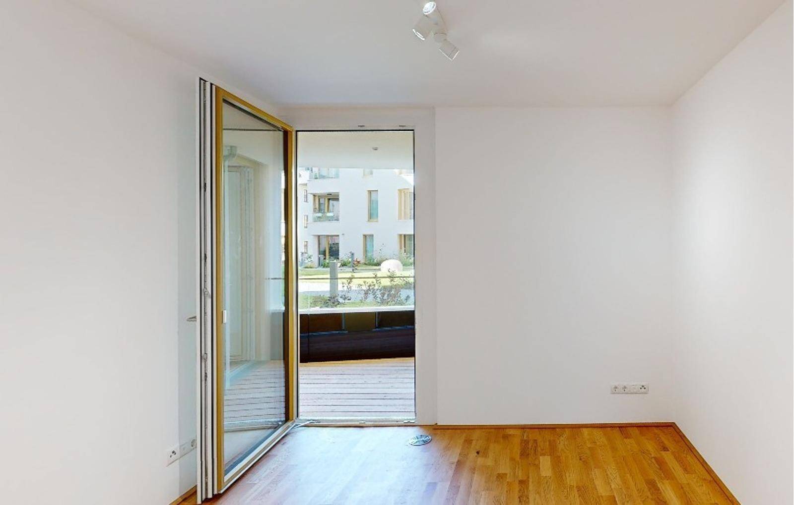 Leerer Raum mit Holzboden und breitem Zugang zu einem Balkon mit Blick ins Grüne.