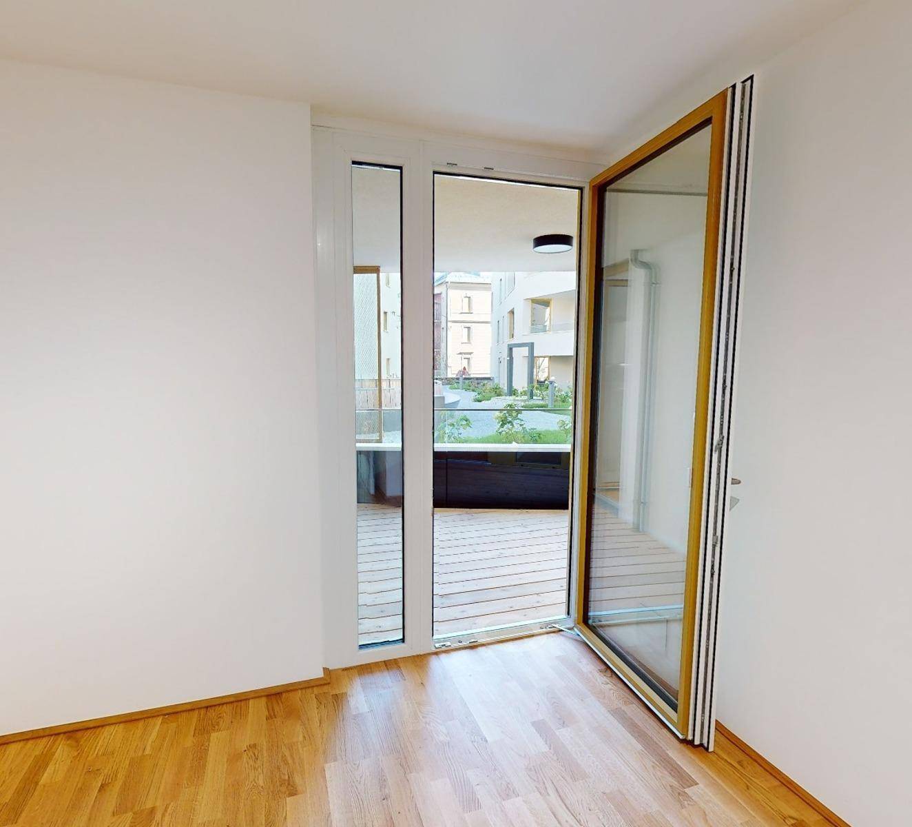 Unmöblierter Raum mit Holzboden und großen Glasfenstern, die einen Zugang zum Außenbereich ermöglichen.